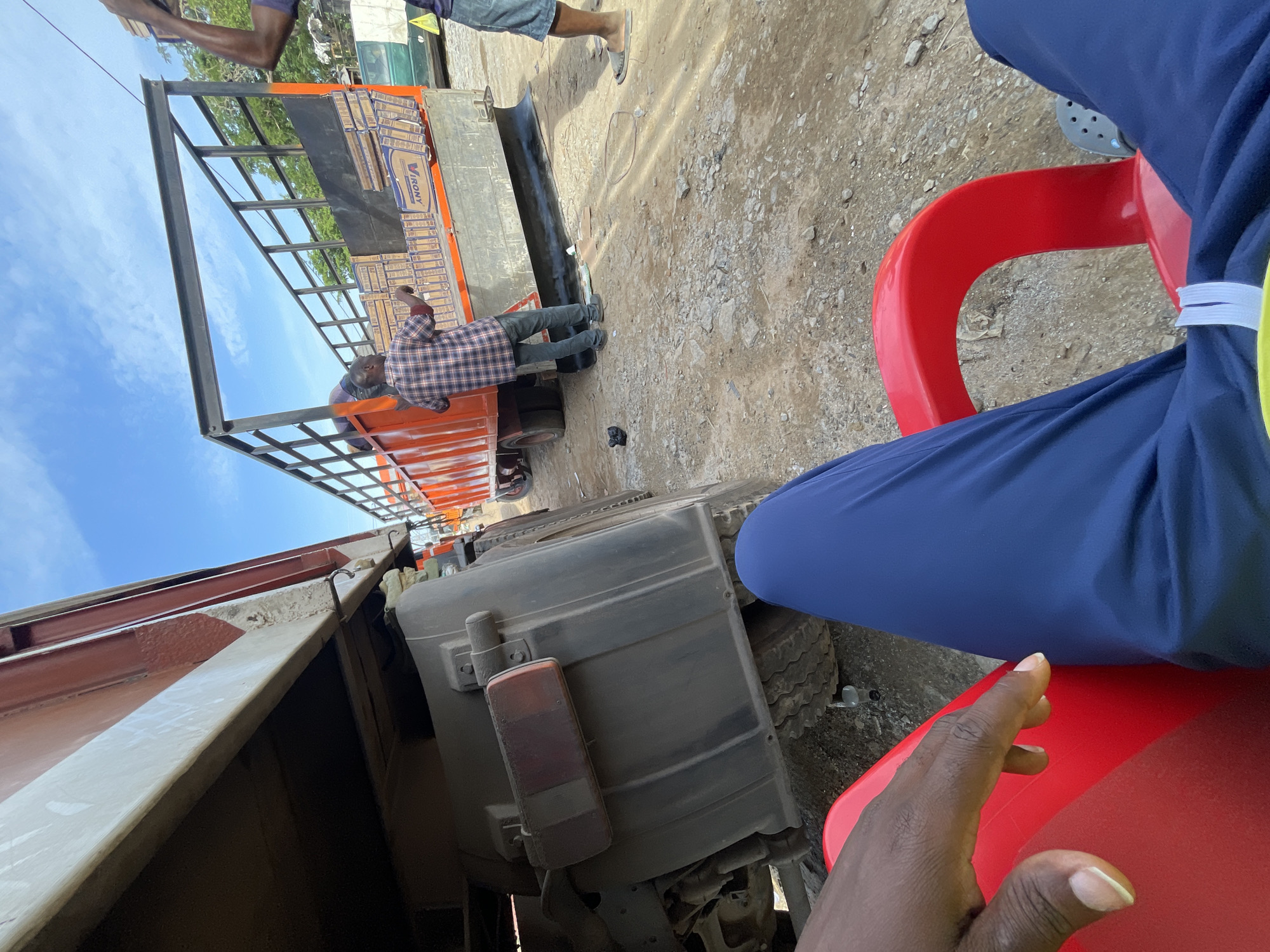 The scene depicts a roadside location in Abuja, Nigeria. A flatbed truck is partially visible, loaded with cardboard boxes bearing the inscription "VIRONIY." A man wearing a plaid shirt stands on the truck bed, presumably involved in loading or unloading the boxes. Another individual, visible only from the knees down, is standing on the ground nearby. The perspective is from someone seated on a red plastic chair, wearing blue pants, and with their hand resting on the chair. The ground is dusty with visible gravel and debris. Parts of other vehicles and structures are also visible. The sky is blue with some clouds.