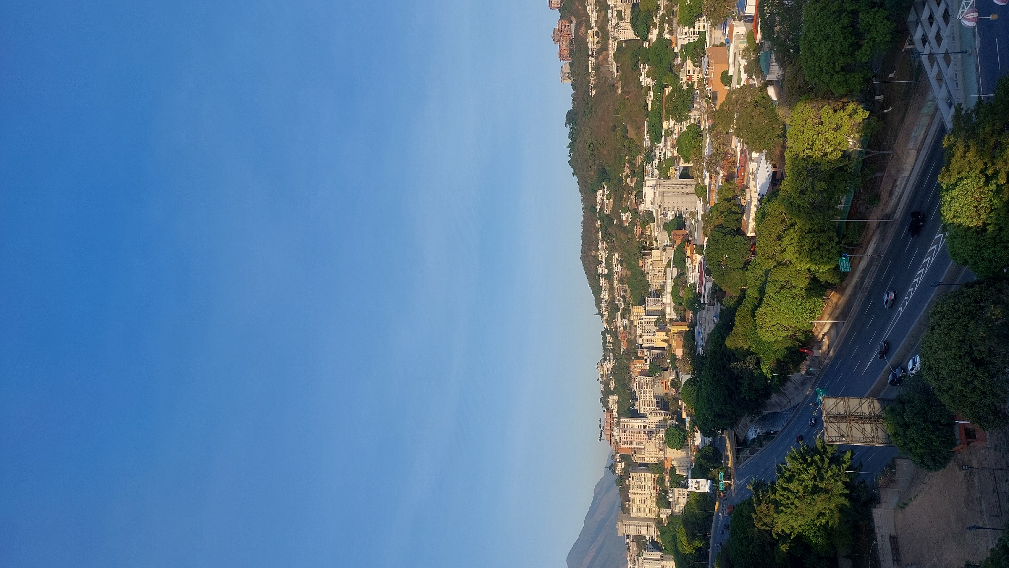 The image shows an aerial view of a city, likely Caracas, Venezuela. The composition is divided between a clear, blue sky on the left and a cityscape on the right. A hillside is densely populated with buildings of varying sizes and architectural styles. A highway runs parallel to the hillside, with several vehicles visible on the road. Trees line the highway and are interspersed among the buildings on the hillside. The overall impression is a densely built urban environment nestled into hilly terrain.
