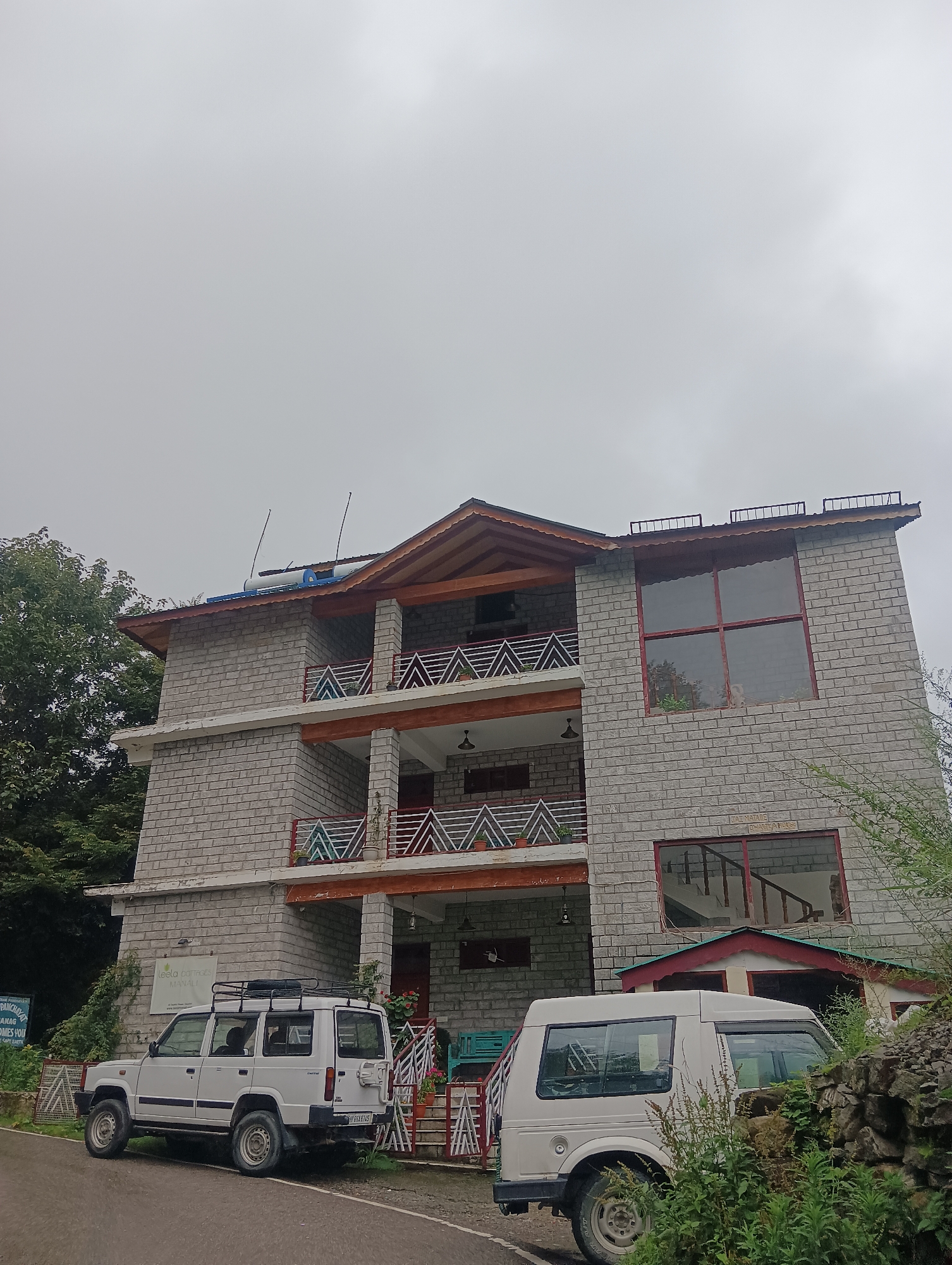 A three-story building made of light-colored brick is situated in Goshal, India. The building has multiple balconies with red railings and decorative white triangular patterns. The roof is sloped and has wooden eaves. Two white vehicles are parked in front of the building on a paved road. The first vehicle, a white SUV, is parked on the left, facing away from the camera. The second vehicle, a white van, is parked on the right, also facing away from the camera. Lush green trees and vegetation surround the building and vehicles, and the sky is overcast. A sign is visible on the building that reads "TEELU COTTAGES MANALI".