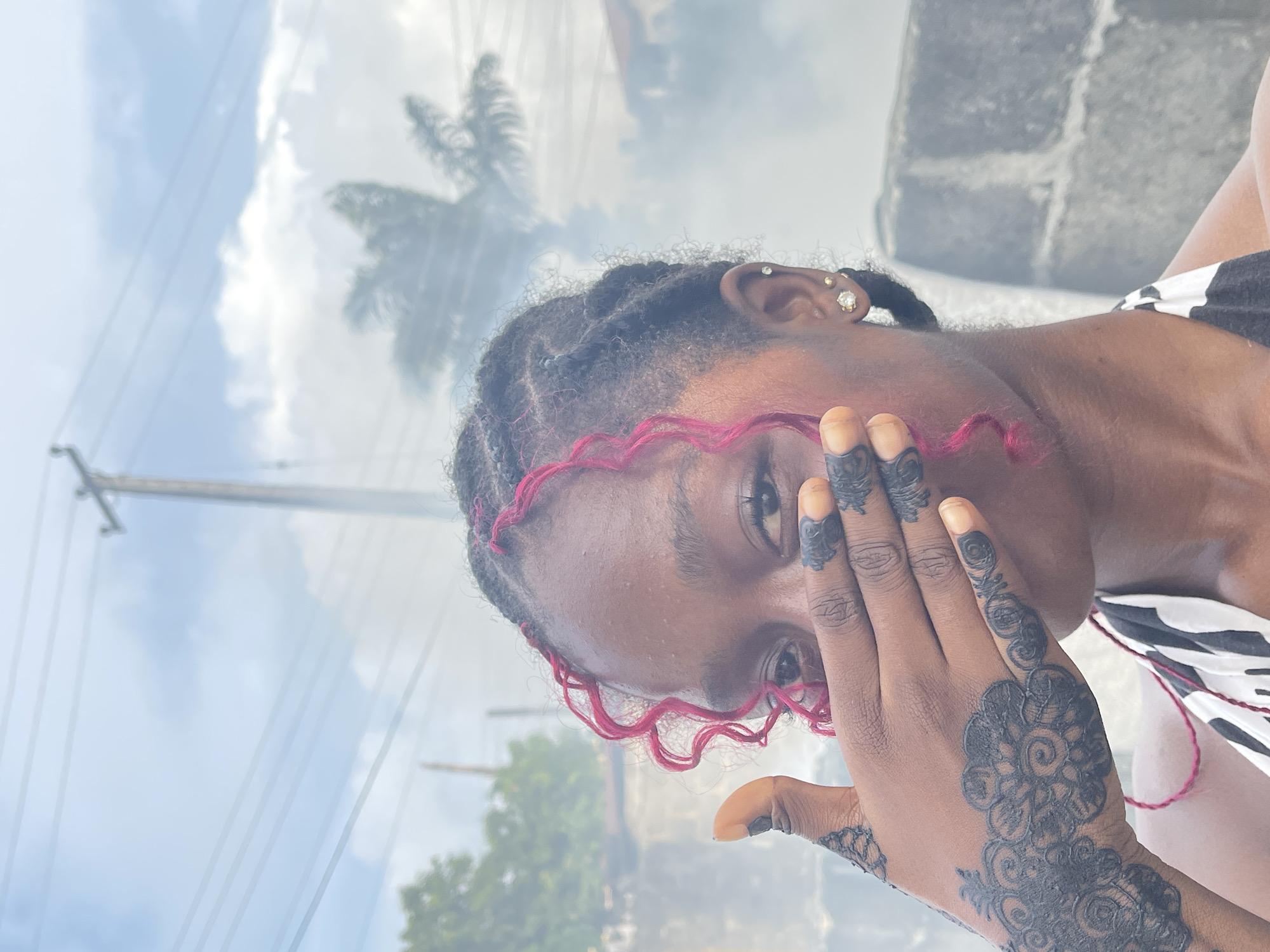 A young, dark-skinned woman poses outside in Ado, Nigeria. She has braids and pink curls framing her face, and is wearing earrings. Her right hand covers part of her face and features intricate henna designs. The background shows a cloudy sky, electrical wires, and a partial view of a palm tree and a building with gray concrete blocks.