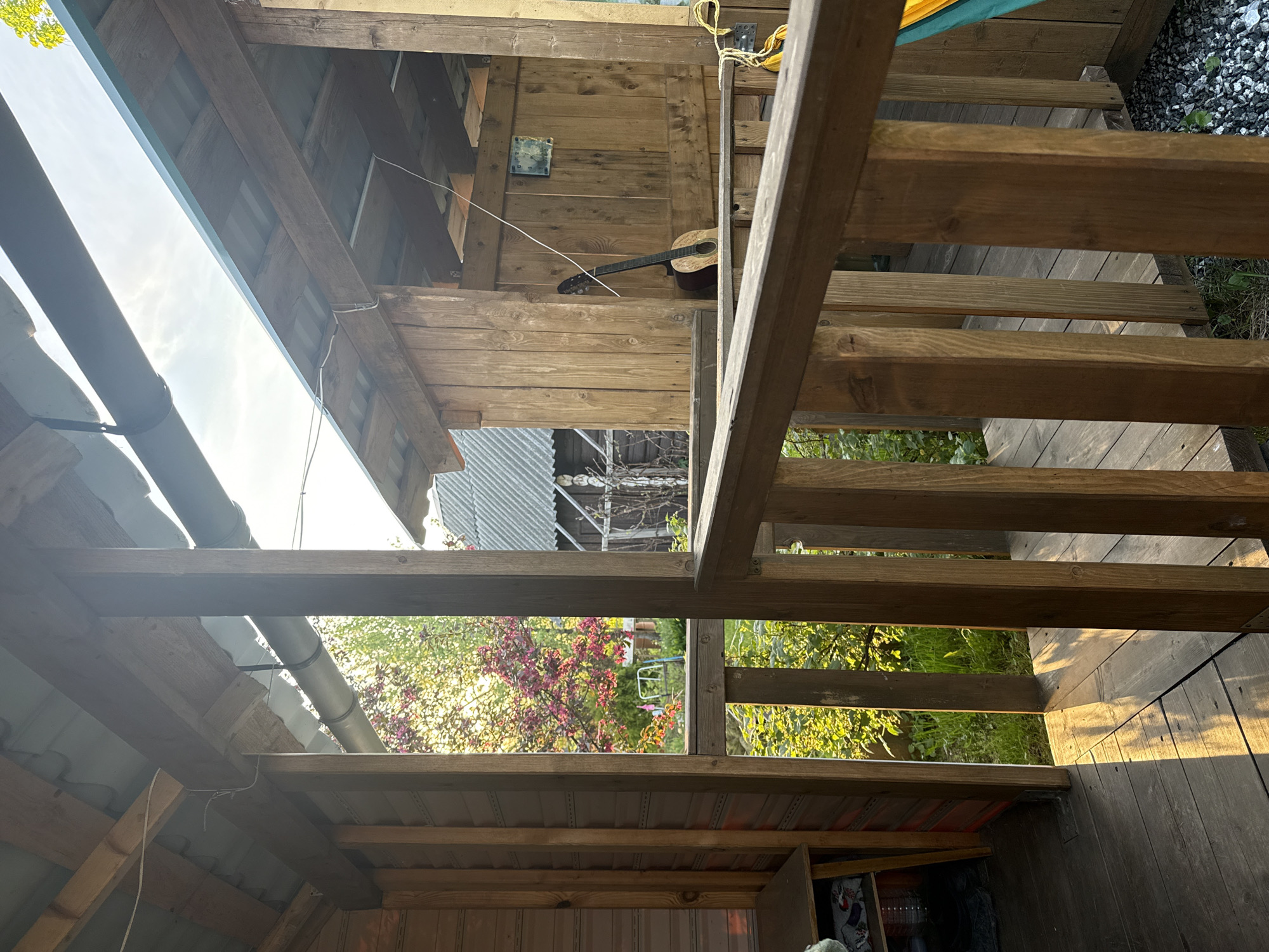 This image depicts an outdoor setting in Kuzili, Russia, possibly within a residential area. The scene is viewed from under a wooden structure, likely a porch or deck, looking out towards a yard. The foreground is dominated by the wooden framework of the structure, including posts, beams, and steps. In the background, greenery is visible, including trees and flowering bushes with pink blooms. A house or building with a corrugated metal roof can also be seen through the framework. An acoustic guitar is leaning against a wooden wall. The lighting suggests either early morning or late afternoon, with sunlight filtering through the trees and casting shadows.