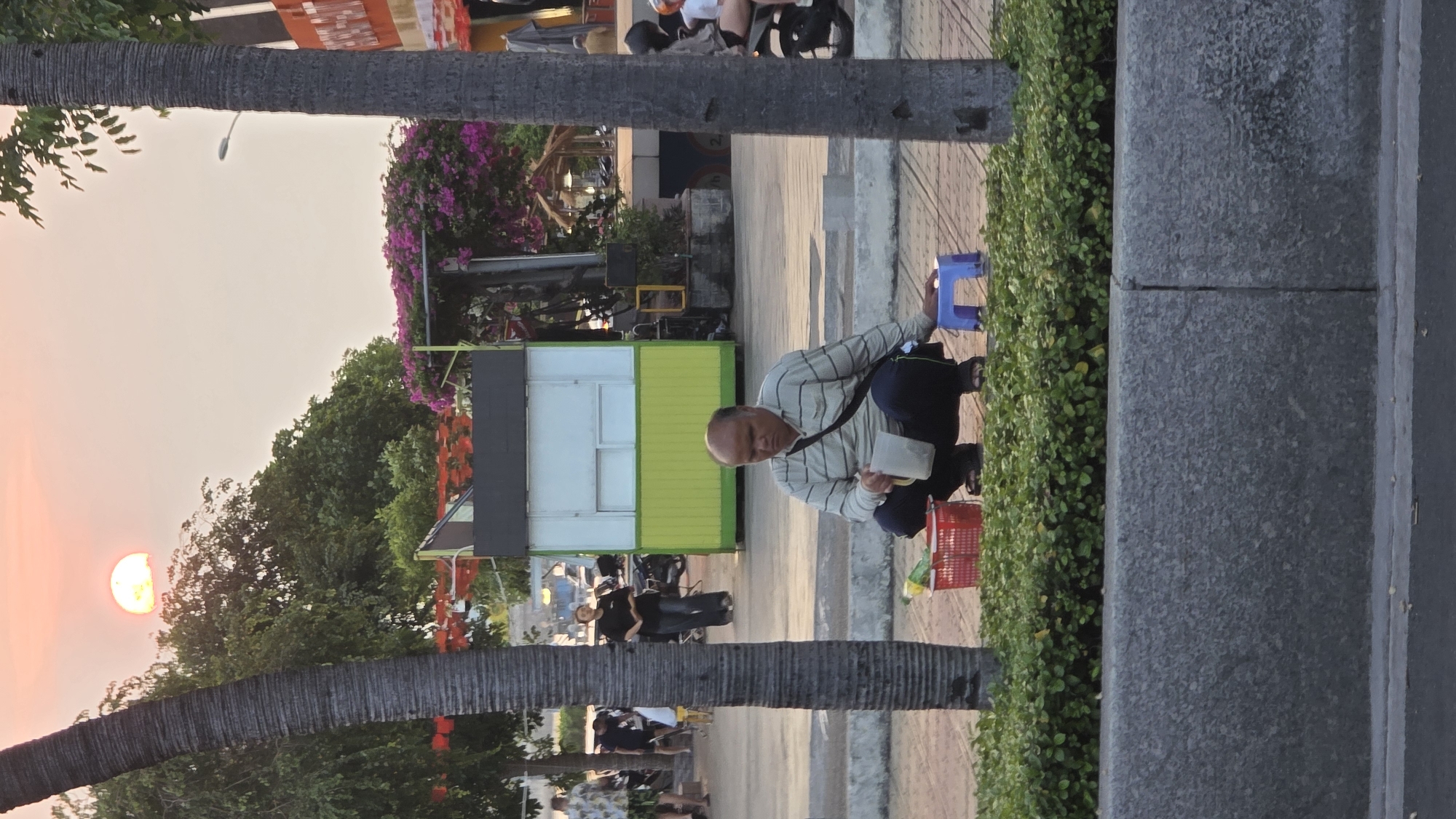 The image, taken at sunset, shows a man squatting on a paved sidewalk in Nha Trang, Vietnam. He is positioned near a green hedge and a low blue plastic stool. He wears a striped shirt, dark pants, and sandals, and carries a bag over his shoulder. He is holding a white object in his left hand. Beside him is a red plastic basket. In the background is a green and white stall, purple flowers, and a palm tree trunk. People and parked motorbikes are visible further down the sidewalk. The sun is setting in the distance, casting an orange glow on the sky.