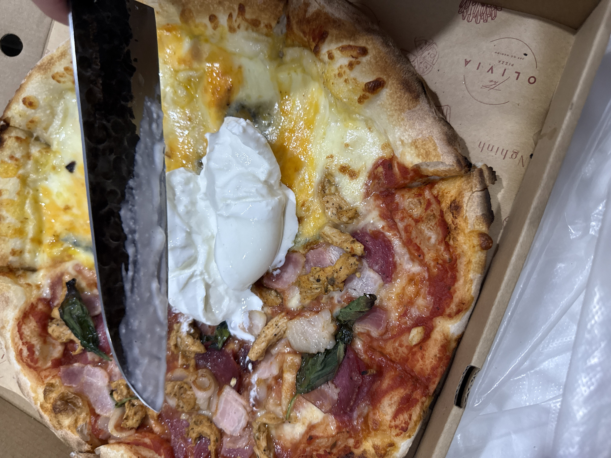 The image shows a partially cut pizza in a cardboard box. The pizza is topped with cheese, tomato sauce, ham, basil leaves, and a large dollop of white creamy sauce. A black-handled knife is cutting through the pizza. The box has the logo "The Olivia Pizza" on it, with the address "305 Ho Nghinh" printed underneath. Ho Nghinh is a street in the city of Đà Nẵng, Vietnam. A white plastic bag lines the box.