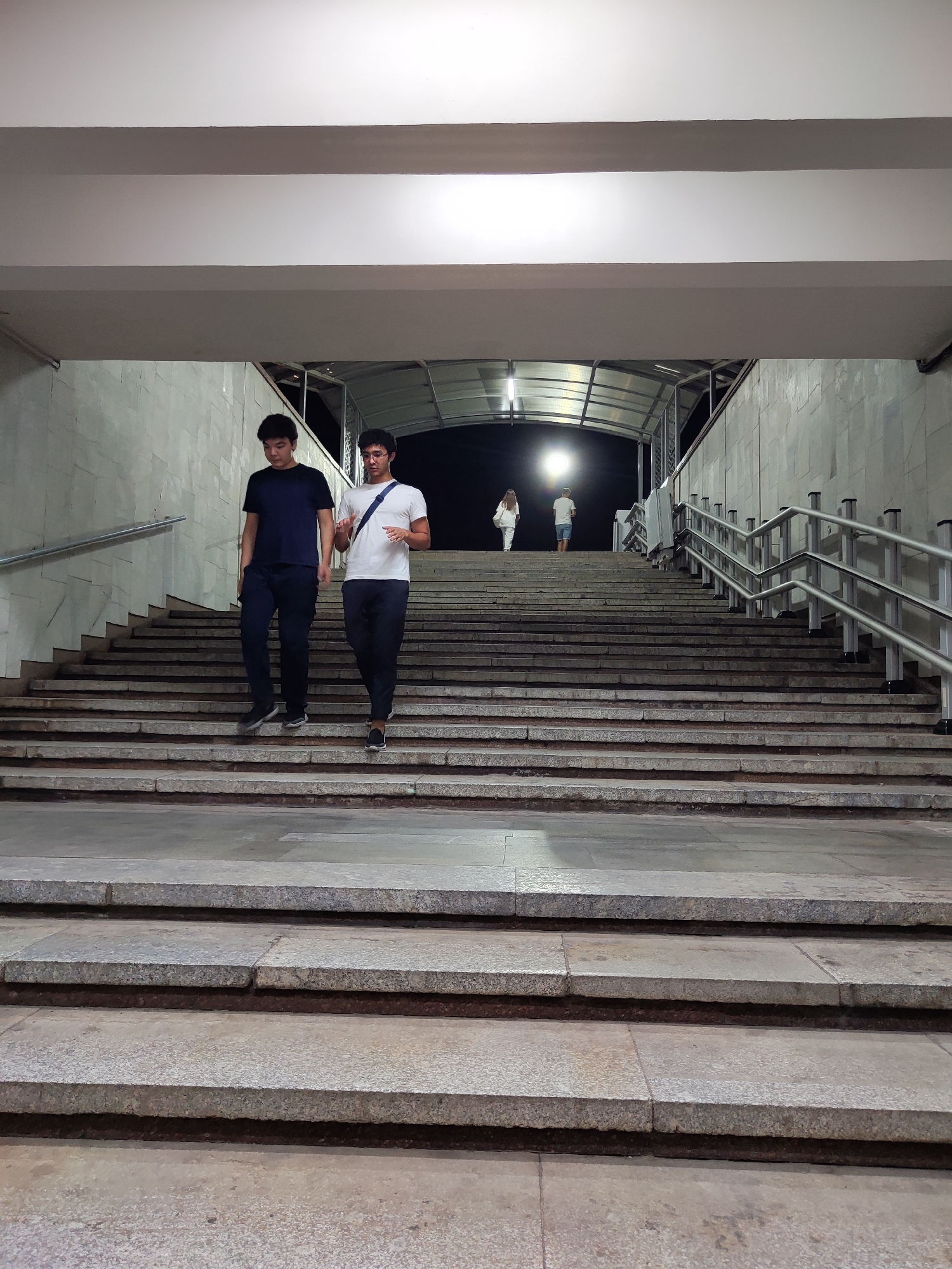 Two young men are walking down a wide, stone staircase in an underpass at night. The man on the left, wearing a dark blue t-shirt and dark pants, is looking down. The man on the right, wearing a white t-shirt with a blue strap across his chest and dark pants, is looking forward and gesturing with his right hand. In the background, two other figures, a man and a woman, are walking up the stairs in the distance under a bright light. The underpass has concrete walls and metal railings along the sides of the staircase. The location is the underpass near the Amir Timur monument in Tashkent, Uzbekistan.