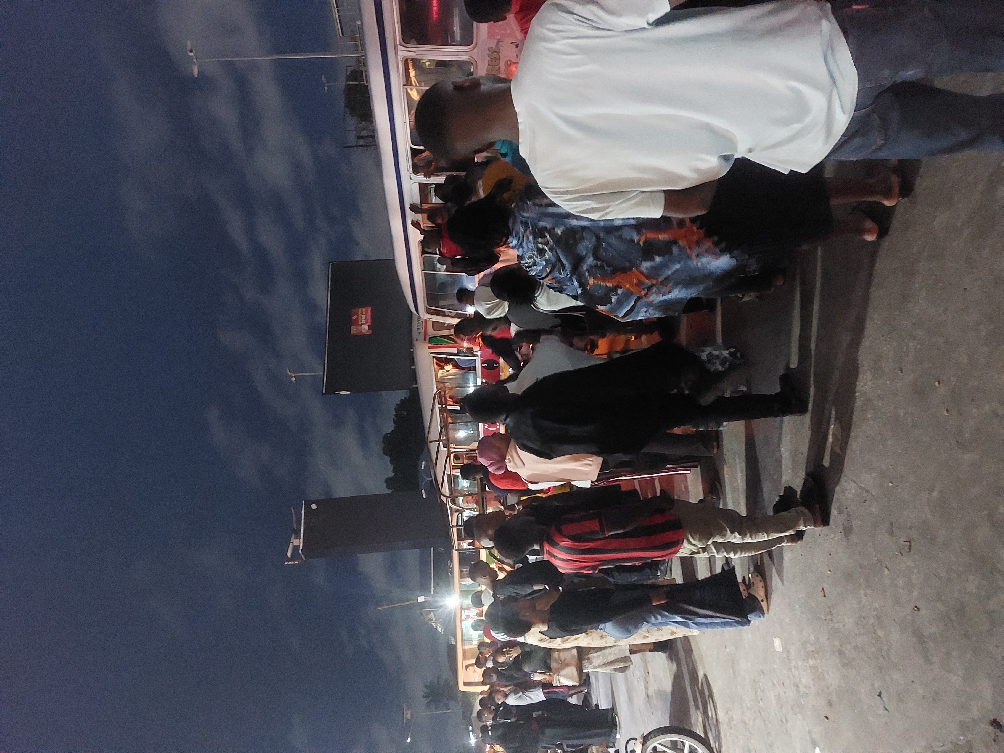 A night scene depicts a bus boarding area with numerous individuals queued up or standing near a bus in Dar es Salaam, Tanzania. People wear casual clothes, including shirts, dresses, pants, and footwear. The bus is white and appears to be full, as some individuals stand on the footboard outside. Street lights illuminate the scene with the dark sky and light clouds overhead. A bicycle and signage are also in the frame. The ground has a rough concrete surface.