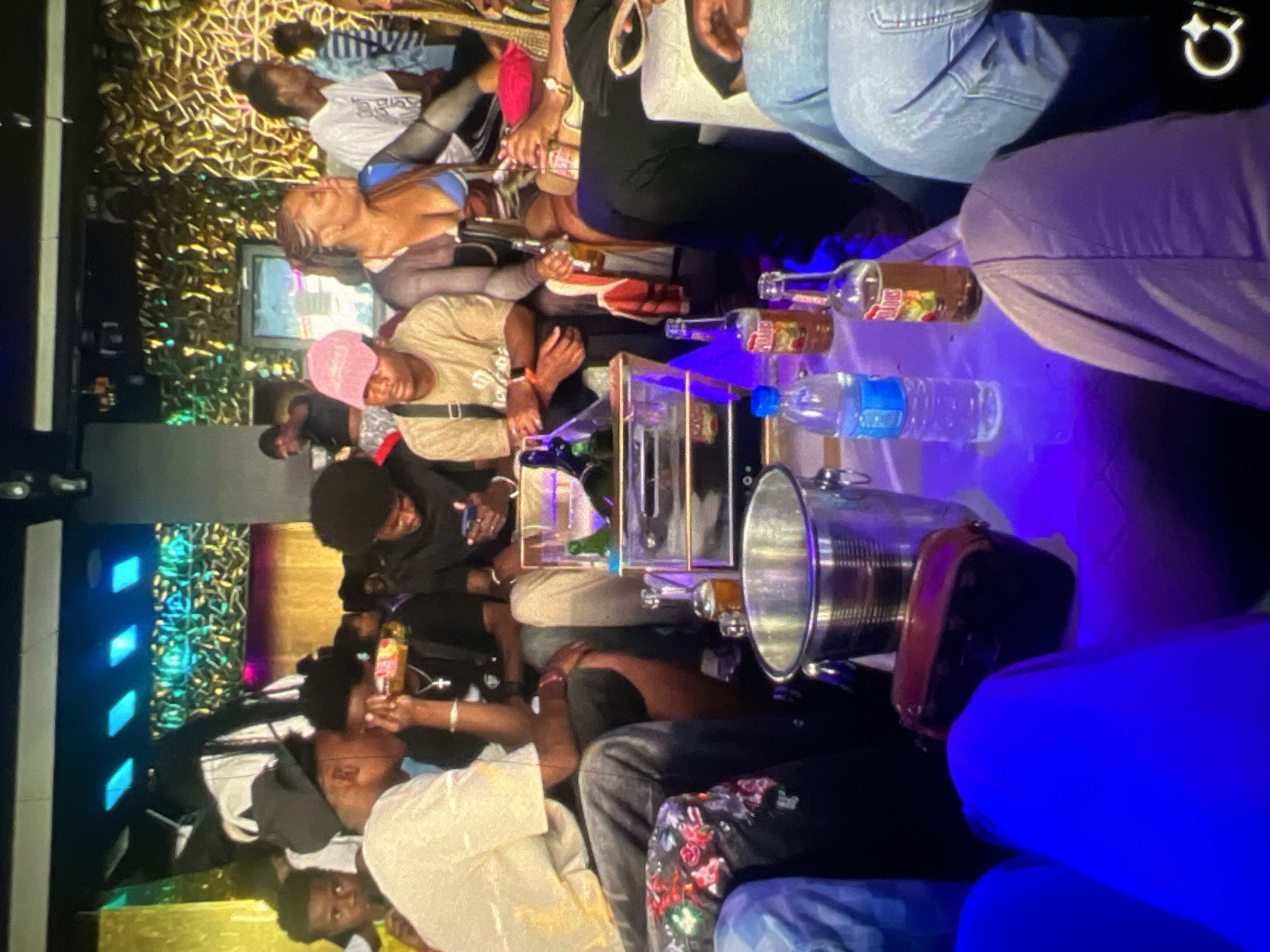 The image depicts a group of young adults gathered around a table in a dimly lit, nightclub-like environment. The setting appears to be a commercial establishment in Lagos, Nigeria, suggested by the context of a vibrant nightlife scene. Several individuals are seated and standing, engaged in conversation and using their mobile phones. A centerpiece on the table includes an ice bucket filled with what appears to be bottles of alcohol and water. Various bottles of alcoholic beverages and a bottle of water are visible on the table, indicating a social gathering with refreshments. The lighting is atmospheric, with blue and purple hues dominating the scene, characteristic of a nightclub or lounge. The individuals are dressed in casual and trendy attire, consistent with a night out. The overall impression is one of a lively social event.