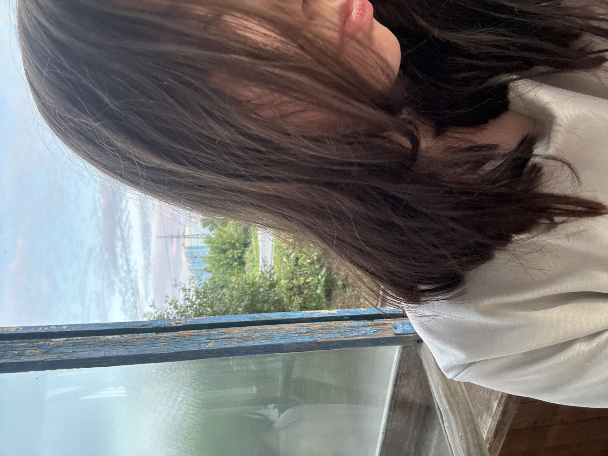 A young woman with long, dark brown hair partially obscuring her face is seen from the chest up, seated indoors. She is wearing a light-colored, possibly white, long-sleeved shirt. The background is a window with a weathered, peeling blue wooden frame, through which trees and a distant cityscape with some industrial structures are visible under a cloudy sky. The windowpane is smudged. The scene appears to be in a rural or semi-rural area, possibly on the outskirts of the city of Inskoy, Russia, suggested by the presence of trees and less dense urban development. The lighting indicates it is likely daytime, perhaps late afternoon or early evening, given the soft, diffused light and the sky's coloration. There is no clear indication of interaction with anyone else.