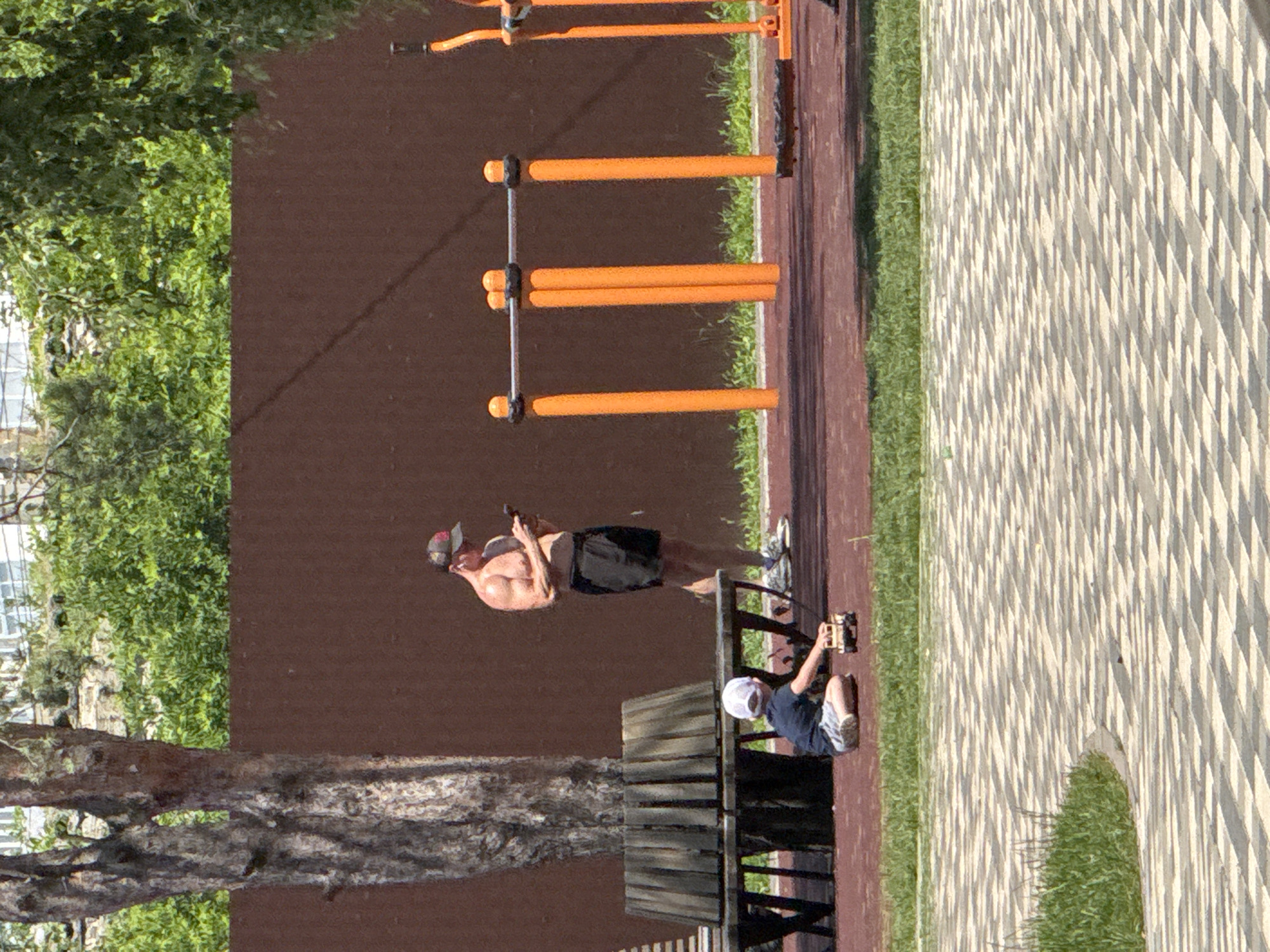In a sunny outdoor setting, a man and a boy are present on a park or playground area equipped with exercise equipment. The man, appearing to be in his late 20s or 30s, is shirtless and wearing dark shorts and a baseball cap. He is holding a smartphone and appears to be engaged with it. The boy, who is younger, is wearing a dark t-shirt, light-colored shorts, and a white baseball cap. He is seated and appears to be interacting with a small object, possibly a toy.
The scene features a series of orange bars, likely part of a pull-up or exercise station, set against a dark brown wall. To the right of the exercise equipment, there is a section of patterned paving stones and green grass. A wooden bench is also visible in the foreground. The lighting suggests it is daytime, with the sun casting shadows. The overall atmosphere seems relaxed and casual, indicative of leisure time spent outdoors. The location is identified as Mikhaylovsk, Russia. There is no visible text in the image.