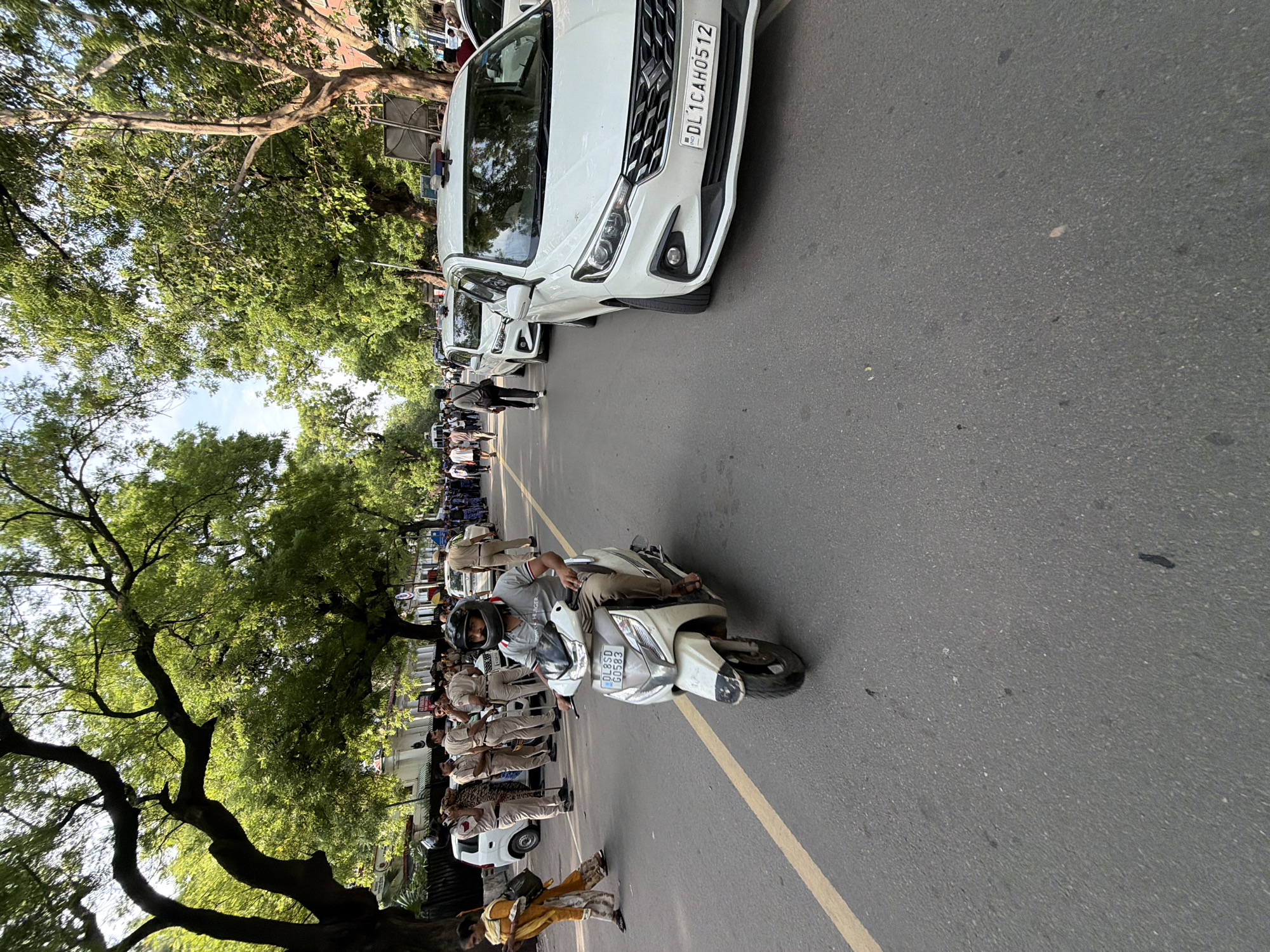 An image shows a street scene in New Delhi, India. The street is paved and a yellow line runs down the center. On the right side of the street, two white SUVs are parked, their lights on. One of the SUVs has a license plate that reads "DL 1 CAH0512". The SUV in the foreground is a Maruti Suzuki Ertiga. On the left side of the street, a man is riding a white scooter. The scooter's license plate reads "DL 8 SD G0583". A woman in a yellow outfit is walking on the left sidewalk. Behind the scooter, a group of men in uniform, likely police officers, are standing. Several trees with green leaves line both sides of the street, and the sky is visible through the branches. The overall scene suggests a street with some official presence or activity.