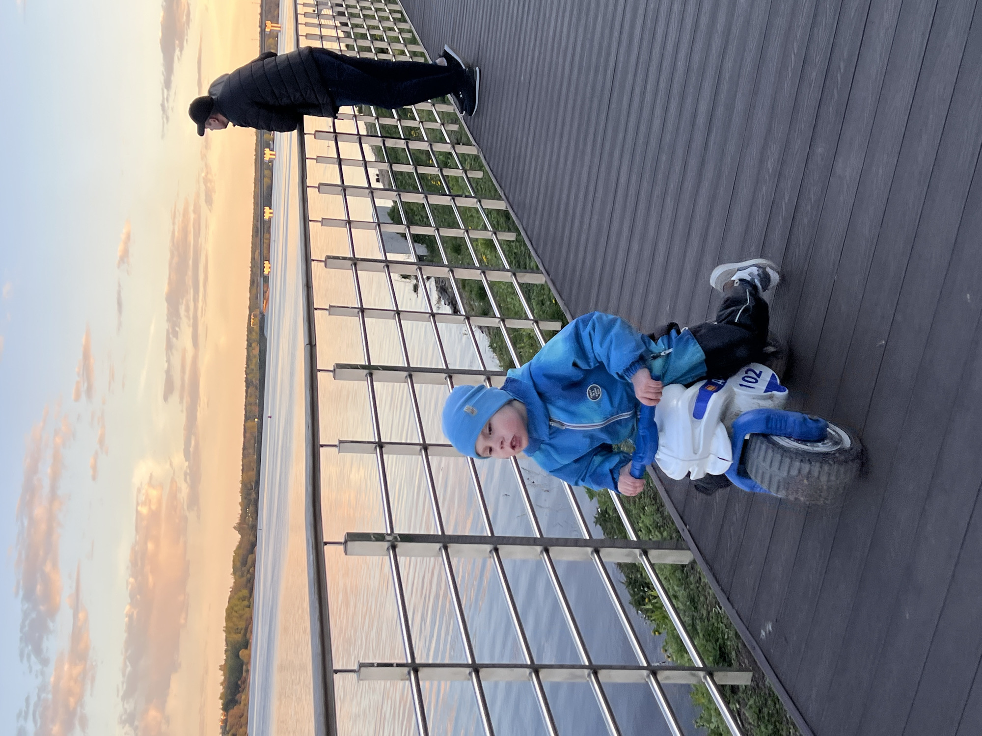 A low-angle, eye-level outdoor shot captures a boy on a toy motorcycle and an adult man on a boardwalk. The scene is set against the backdrop of the Sheksna River in Cherepovets, Russia, as suggested by the distinctive shoreline and cityscape visible in the distance.
The boy, wearing a bright blue jacket and matching beanie, straddles a white and blue toy motorcycle. The toy's tires are visibly worn, and the number "102" is visible on the motorcycle. He is positioned near a silver metal railing.
In the background, an adult man in a black jacket and black trousers stands leaning over the railing, looking out at the river. The boardwalk is made of dark-colored wood planks that run diagonally through the frame. The river reflects the sky, which is painted with the colors of dawn or dusk. The presence of trees lining the opposite bank suggests a natural element intermingling with the urban setting.