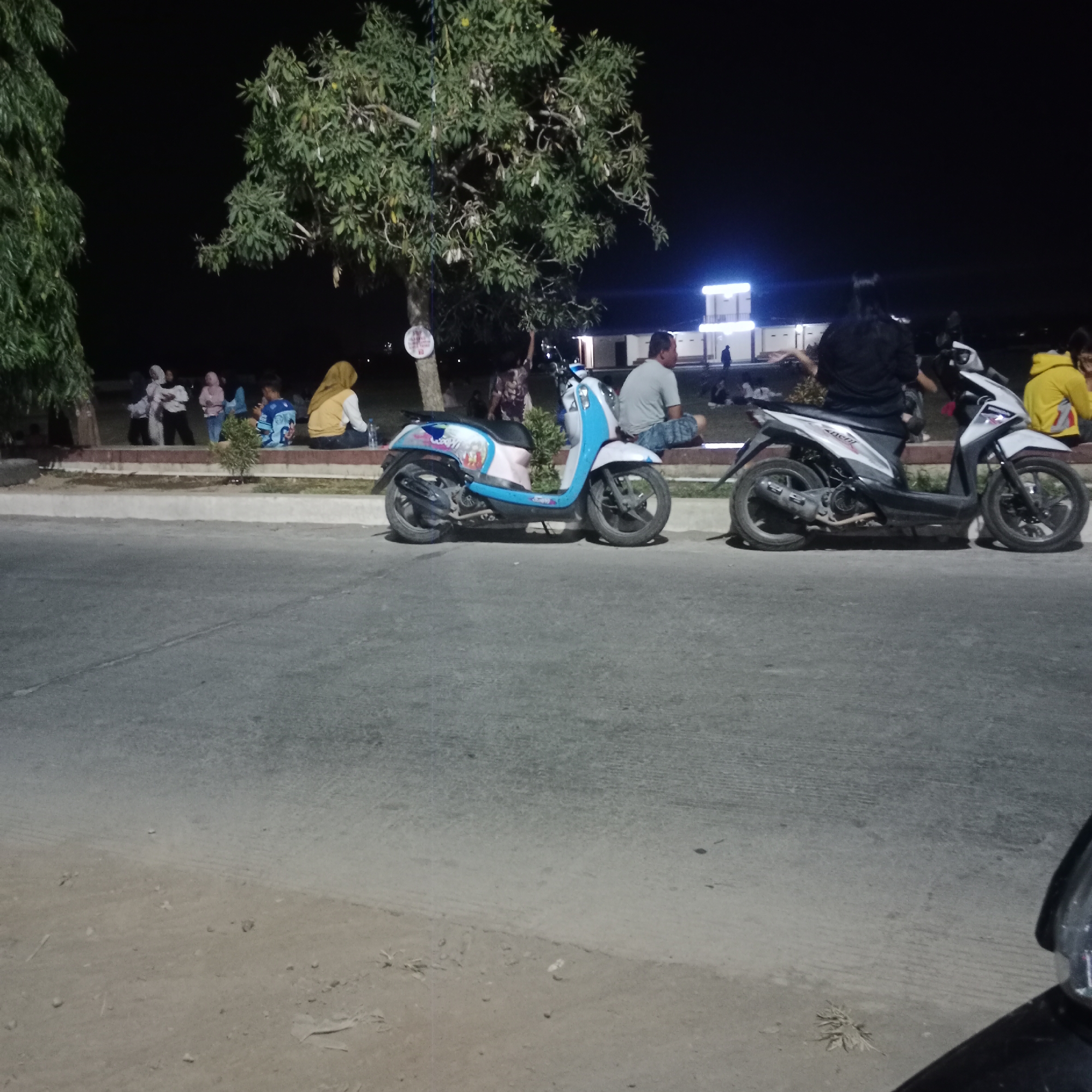 The image depicts an outdoor scene at night, likely in Kunjeng, Indonesia, as indicated by external information. In the foreground, a paved road is visible, with a dirt and gravel embankment running alongside it. Two scooters are parked on the embankment. The scooter on the left is predominantly light blue and pink with decals. The scooter on the right is white and black.
Behind the scooters, a group of people are gathered, some sitting and some standing. They are situated on a raised, paved area, possibly a sidewalk or seating area, next to a grassy strip. A large tree with green foliage is prominent on the left side of this area, and another tree is visible further back. In the background, several buildings are illuminated by bright lights, including a structure with a prominent white facade and blue lighting. The sky is dark, indicating nighttime.
Among the people visible, several are wearing headscarves, suggesting a Muslim-majority population. One person in a bright yellow jacket is seated on the right. Another person in a black shirt is standing and appears to be interacting with a scooter. The overall atmosphere appears to be relaxed, with people congregating outdoors in the evening. The presence of scooters suggests this is a common mode of transportation in the area. The illuminated buildings in the background might be commercial establishments or public facilities.