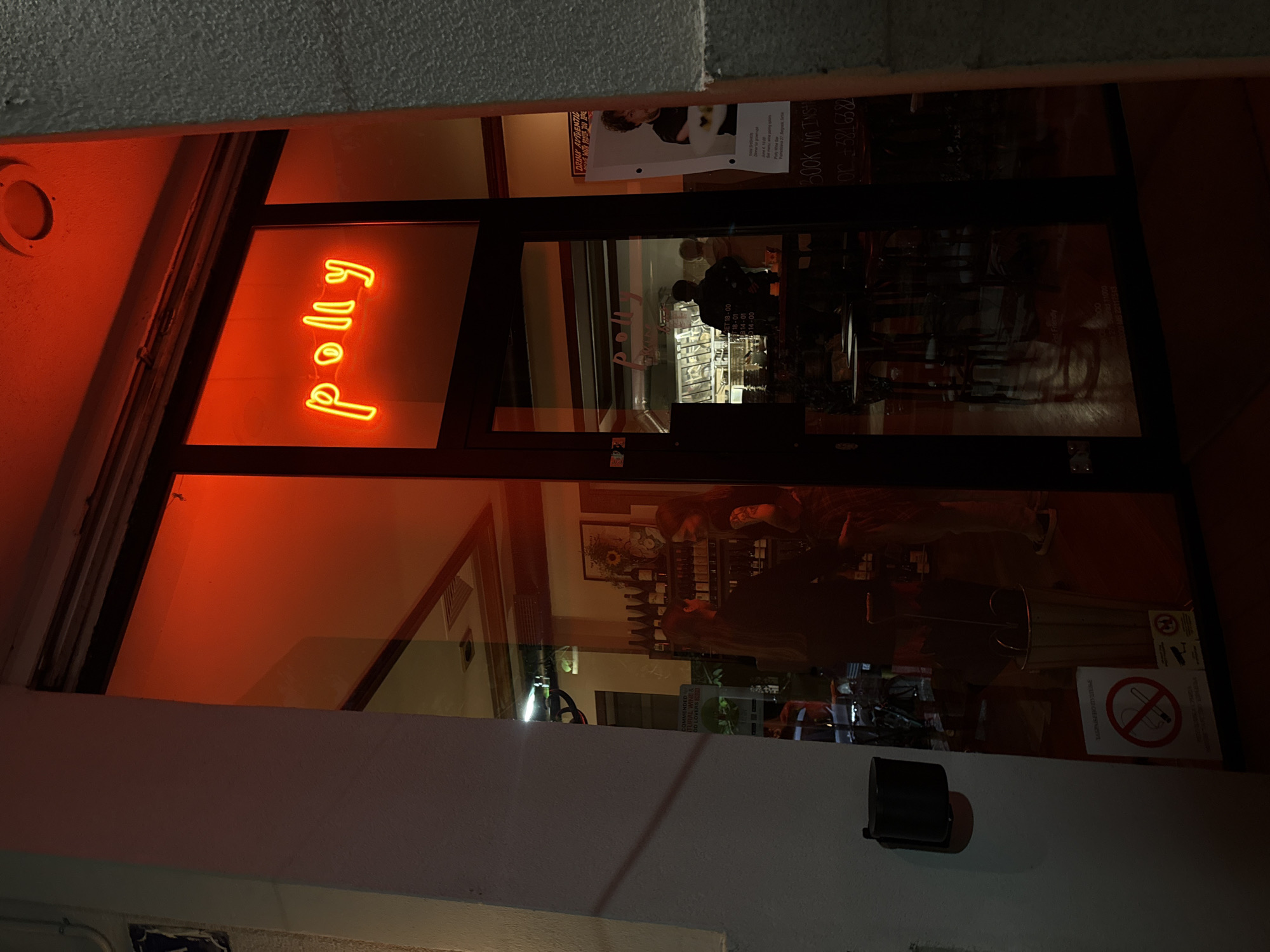 The image captures the glass storefront of an establishment, identified by a neon sign as "polly," at night in Belgrade, Serbia. The scene is dominated by the warm, intense reddish-orange glow emanating from the vertical neon sign and reflecting on the surrounding surfaces.
Through the glass, the dimly lit interior of what appears to be a wine bar or shop is visible, along with reflections from both inside and outside. Inside, shelves stocked with bottles, likely wine, are discernible. Blurred figures of people are present; one person with dark hair and a light shirt is visible behind a counter in the upper part of the doorway, and another partial figure is seen in the lower right reflection.
Various signs and posters are affixed to the glass or inside. A prominent red and white circular sign with Cyrillic text reading "ЗАБРАЊЕНО ПУШЕЊЕ" (No Smoking) and its English translation "NO SMOKING" is visible. Other text includes "COMMENDED NATURAL WINE & GOOD LOVERS," "DRINK AUTHENTIC," and information about an event: "June 4, 18:00," at "Polly Wine Bar, Belgrade, Serbia." Contact details "Book via Inst" and "or +3816382" (a phone number) are also present. A list of numbers like "OPEN 18-00, 18-01, 314-01, D14-00" suggests operating hours or internal codes.
The rough, textured exterior wall of the building is visible on the left and top, and a dark, wall-mounted fixture is located below the right side of the doorway. The overall ambiance is dark and inviting, typical of an evening scene at a bar or cozy establishment.