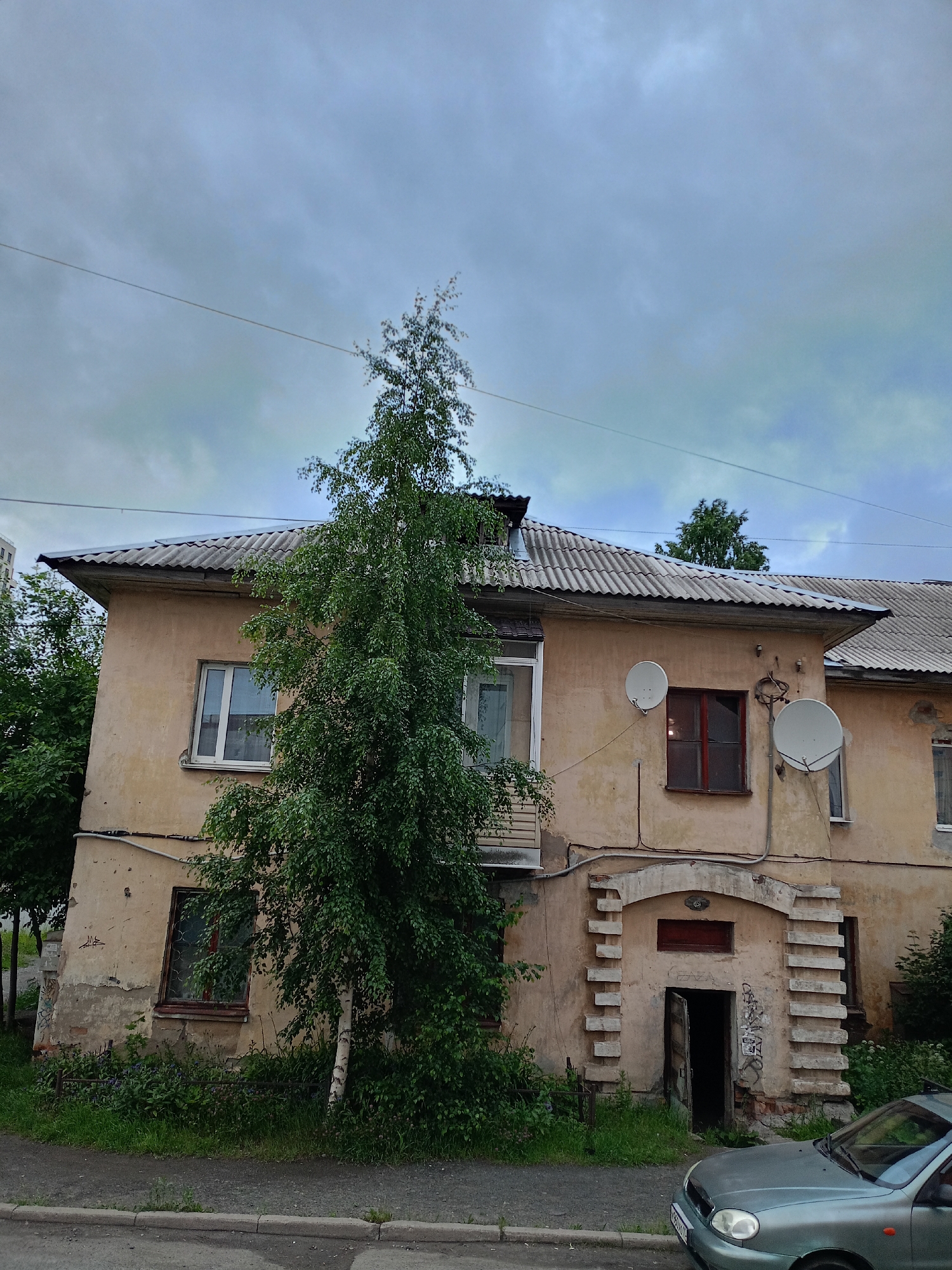 The image shows the exterior of a two-story, light-yellow apartment building with a corrugated metal roof. A tall, slender birch tree with lush green foliage is positioned in front of the building, obscuring a portion of its facade. The building has several windows, some with red frames and others with white frames. Two satellite dishes are mounted on the exterior wall. There is an arched entryway with decorative brickwork leading to an open door, with graffiti visible on the wall next to it. A portion of a light blue car is visible in the lower right corner of the image. The sky is overcast with gray clouds. The building appears to be located on a street with a sidewalk and a road. The context provided by the user indicates the location is the city of Petrozavodsk, Russia.