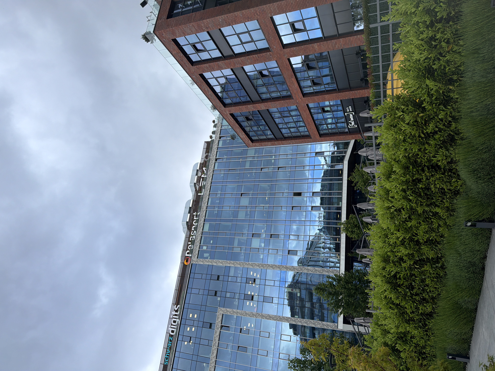 The image displays a modern urban landscape featuring two distinct architectural styles under an overcast sky. A prominent multi-story building occupies the left and central parts of the frame, characterized by its reflective blue glass facade that mirrors the cloudy sky and adjacent structures. Several windows on this building are open or tilted. Visible signage on its upper section includes "Schwarz digiits" in white lettering and "De'a_sport" with a circular, colorful logo.
To the right, a brick-clad building rises, featuring large, dark-framed square windows, some of which are also open. This brick structure appears to cantilever over the lower glass building. At its base, a sign for "Fuel & Water - bar & snacks" is visible, suggesting a commercial establishment.
The foreground and bottom right of the image showcase meticulously maintained landscaping, with rows of vibrant green bushes and neatly trimmed grass bordering a light-colored paved area. Partially visible amidst the greenery and beneath the buildings are several outdoor umbrellas and tables, indicating an outdoor seating area. No people are present in the scene. The setting appears to be a contemporary commercial or office complex, under a grey, cloudy daytime sky. The location is Sofia, Bulgaria.