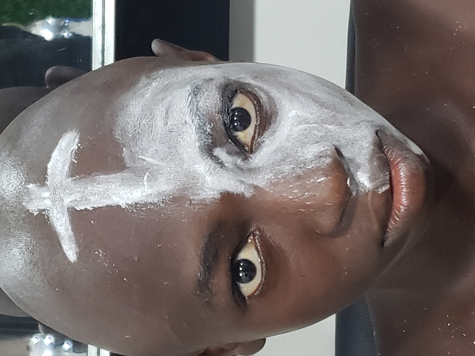 A close-up, angled portrait shows an individual, likely a child or young person, with dark skin. Their face is adorned with white paint, creating striking patterns. A prominent white cross symbol is painted on the forehead, and white substance also covers areas around the eyes, nose, and cheeks. The person's wide, dark eyes look directly forward, conveying a neutral or curious expression. Their lips are visible, appearing natural and slightly parted. A subtle sheen is present on the skin, possibly due to sweat, oil, or the nature of the paint. Parts of the person's shoulder or chest are visible on the right, also showing sparse white specks, suggesting the paint extends beyond the face. The background is out of focus and indistinct, showing a mix of dark and light tones with some reflections, suggesting an indoor or shaded environment. This imagery likely depicts a cultural event, ritual, or celebration specific to Adankolo, Nigeria, where face painting is a common practice. No visible text is present in the image.