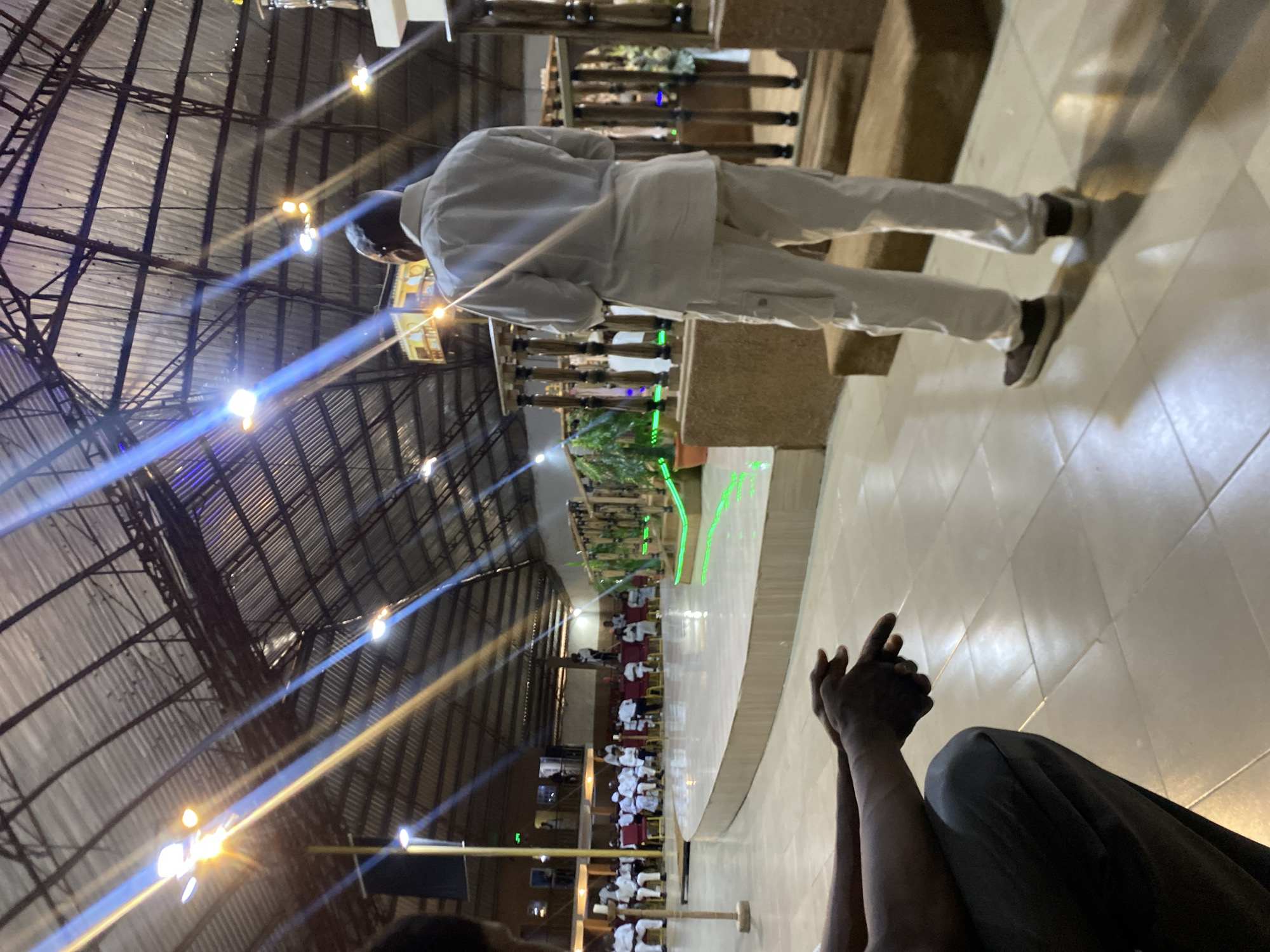 A person in a white shirt and pants stands near a railing in a large, indoor space, possibly a church or auditorium on Rukuba Road, Nigeria. The ceiling is visible, with metal beams and a corrugated metal roof, and numerous lights cast a bright glow. There is a crowd of people seated in rows below the person by the railing. In the foreground, two dark-skinned hands are clasped together, indicating someone is sitting and observing the scene. The floor is tiled.