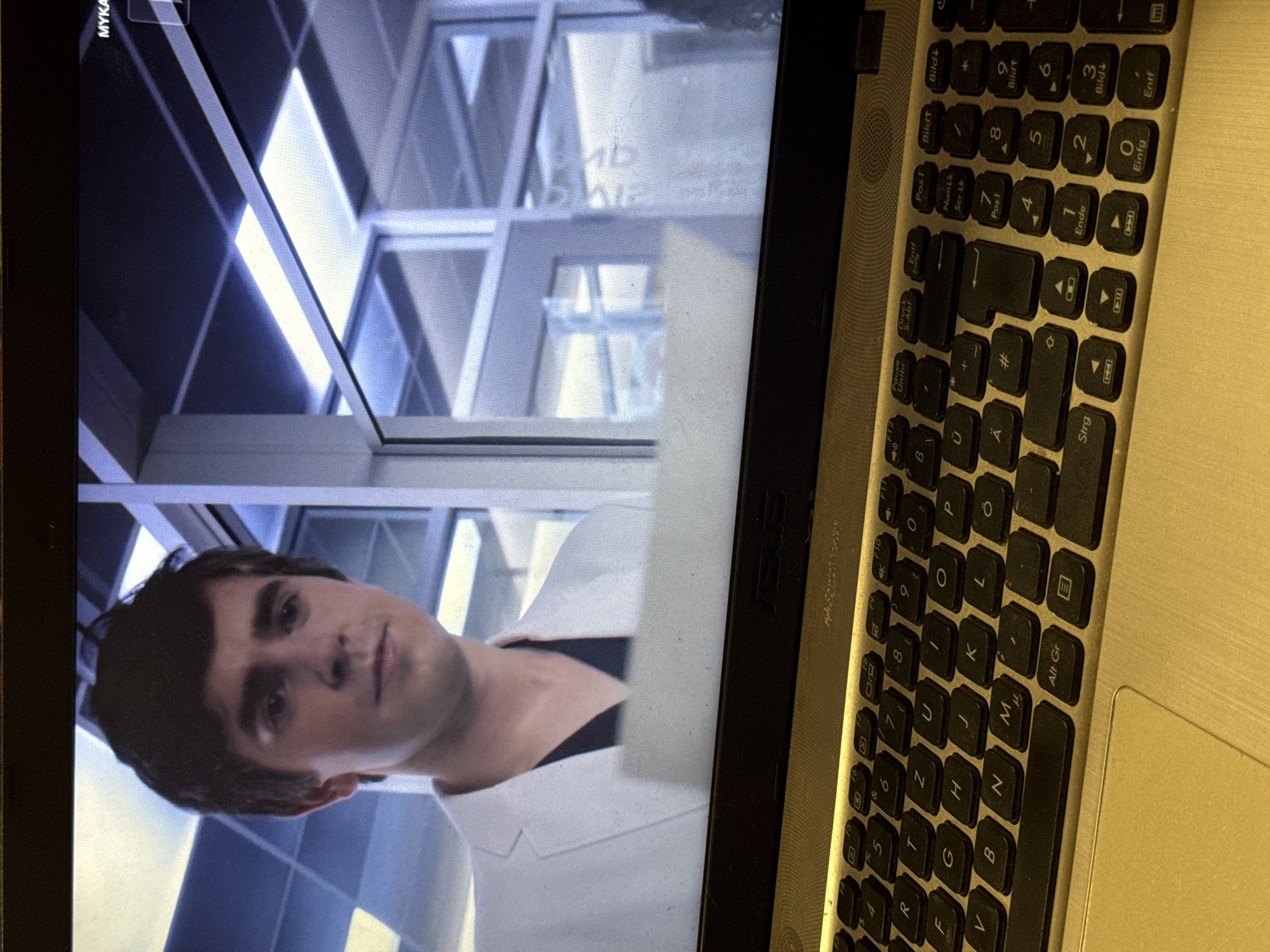 The image shows a close-up view of a laptop keyboard in the foreground, with a television screen displaying a scene from a medical drama in the background. The television screen features a young man with dark hair, wearing a white lab coat over a dark V-neck shirt. He appears to be a doctor, with a serious expression on his face. The background on the screen shows a modern hospital interior with large glass doors and windows, and fluorescent lighting. The laptop keyboard is black with white lettering, and some keys are illuminated. The overall scene suggests a person is watching television while using a laptop. The location is unknown.