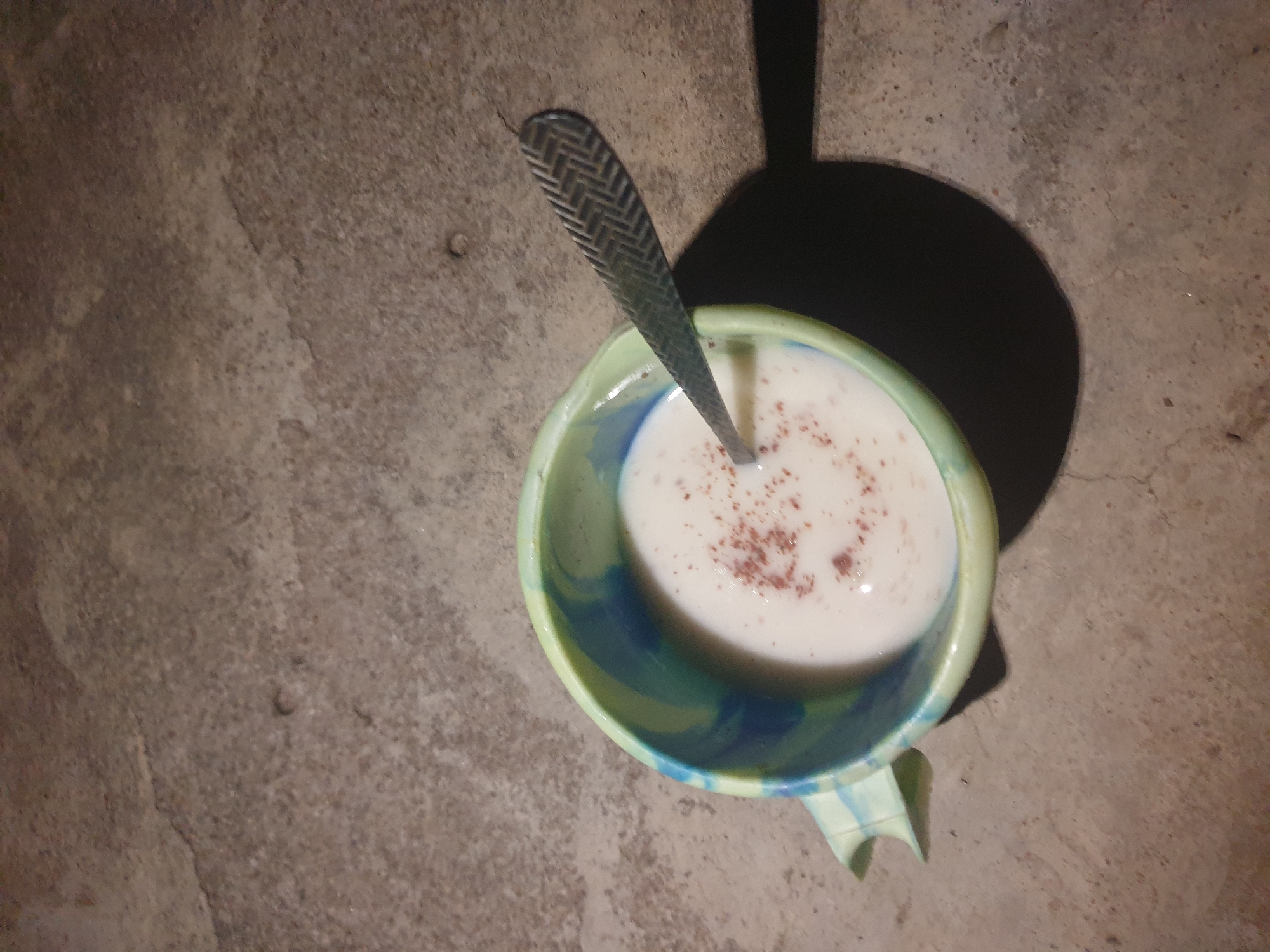 The image shows a close-up, high-angle view of a blue and green swirled ceramic bowl filled with a white liquid and sprinkled with dark brown powder. A patterned metal spoon rests in the bowl, partially submerged in the liquid. The bowl and spoon are situated on a rough, textured concrete surface. A dark shadow is cast to the right of the bowl, indicating a light source from the upper left. The scene is set in Jalingo, Nigeria, where such bowls and concrete surfaces are common.