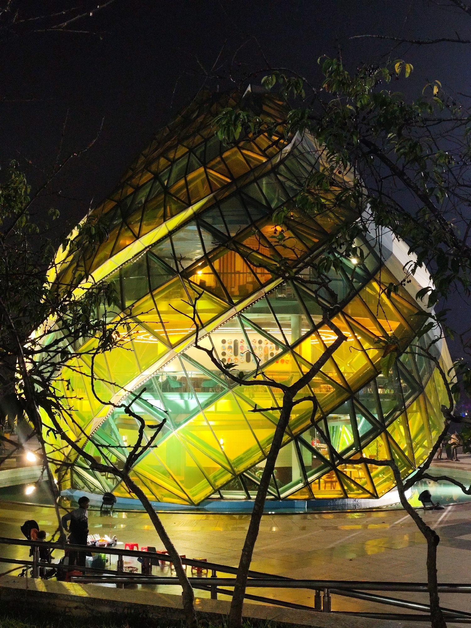 This nighttime photograph captures a striking, geometrically-patterned building in Da Lat, Vietnam. The structure is composed of numerous triangular and rectangular glass panels, illuminated from within by vibrant yellow and green lights, giving it a multifaceted, jewel-like appearance that reflects its surroundings.
In the foreground, the dark, gnarled branches of a tree, some with sparse foliage, frame the brightly lit building. The ground below, likely pavement or concrete, appears wet, reflecting the building's vivid colors and suggesting recent rain or damp conditions.
On the lower left side of the frame, an outdoor vendor stall is set up. A person stands behind the stall, attending to items displayed on a table, possibly food or merchandise, alongside several small red plastic stools. Another individual is partially visible nearby, possibly with a stroller. A dark metal railing runs across the mid-ground, separating the viewer from the vendor area.
The sky is a deep black, emphasizing the artificial illumination of the building and the vendor's setup, creating a strong contrast. The overall scene depicts a lively, urban evening atmosphere. No discernible text is visible within the image.