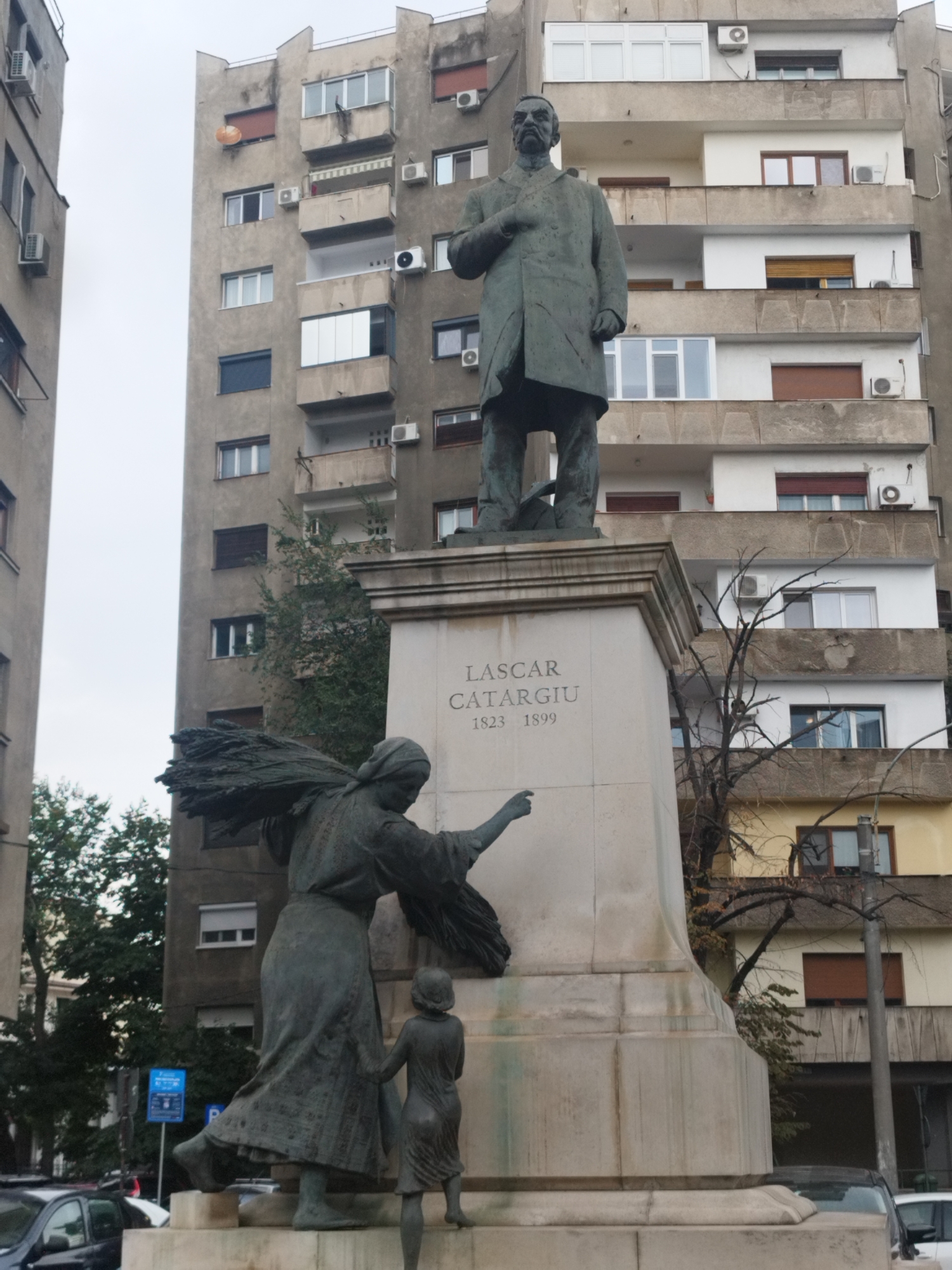 In Bucharest, Romania, a monument stands before a utilitarian apartment building. The monument features a large bronze statue of a man, dressed in a long coat and standing with his hand on his chest. In front of him, a smaller bronze statue depicts a woman carrying a sheaf of wheat over her shoulder and holding the hand of a child. The pedestal of the monument is inscribed with the name "LASSCAR CATARGHIU" and dates "1823 1899". Behind the monument, the grey facade of a multi-story apartment building with numerous windows and balconies is visible. Trees with bare branches are also present, adding to the urban landscape.