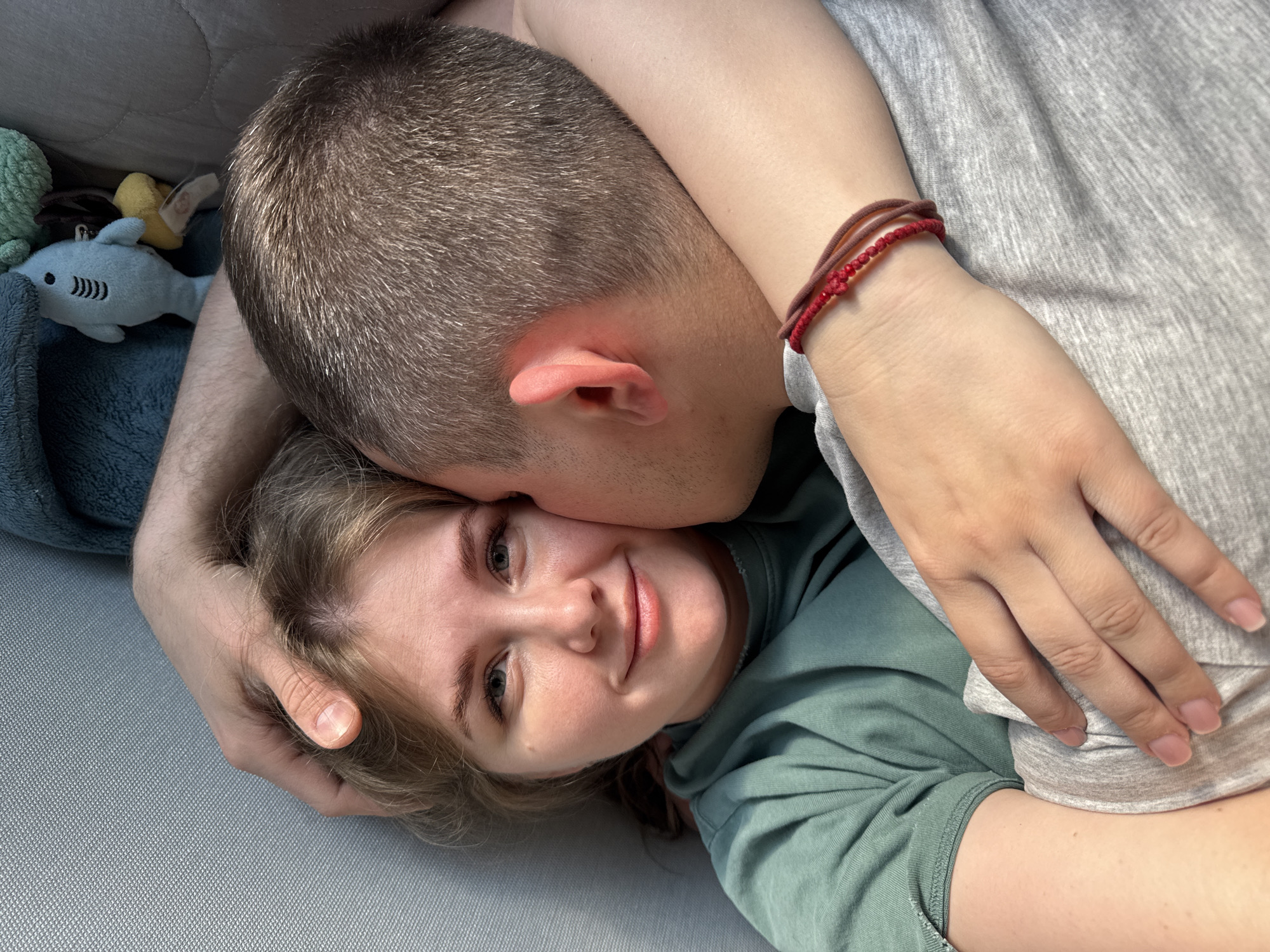 A couple is lying on a gray surface, possibly a sofa or bed. The woman, with fair skin and light brown hair, is facing the camera, smiling. She is wearing a green shirt. A man, with short brown hair, is positioned behind her, with his arm around her head. He appears to be kissing her cheek or whispering something in her ear. He is wearing a gray shirt and has a red and brown bracelet on his wrist. To the left of the couple, there is a blue plush shark and other indistinct plush objects. The scene appears to be indoors, likely in a home. There is no indication of the location being Ферма, Russia.