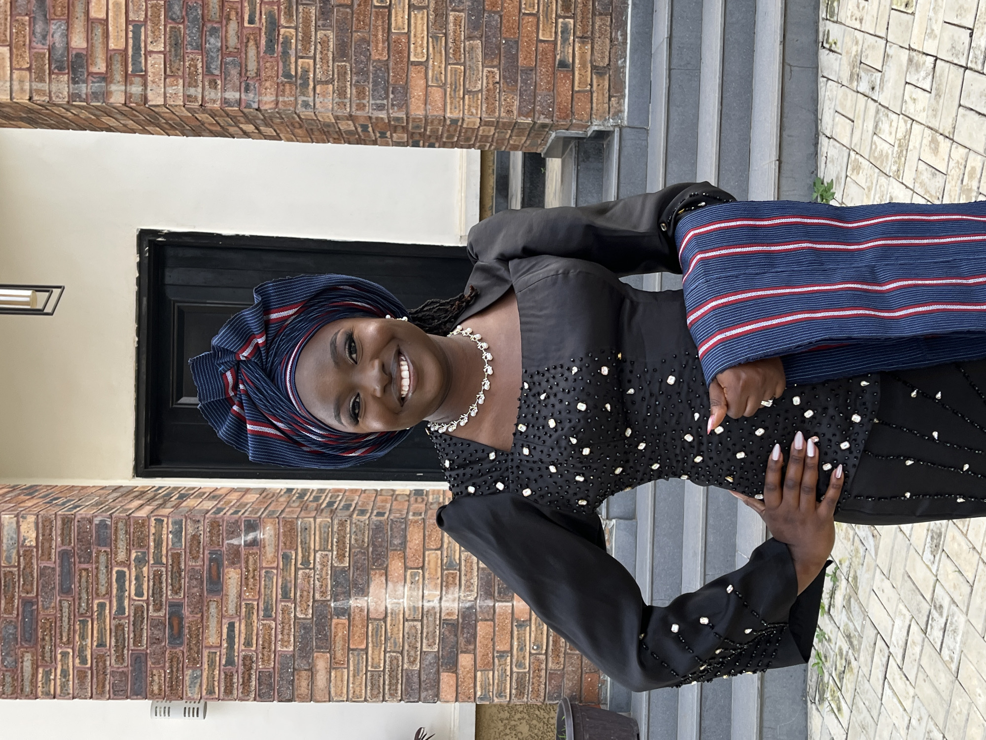A Black woman stands outside a building in Gura Top, Nigeria, wearing a black embellished top with puffed sleeves, a white beaded necklace, and a blue, white, and red striped headwrap and matching shawl. She is smiling and looking at the camera. The building behind her is made of red brick and has a black door and a set of grey stairs. The ground is paved with interlocking bricks. The scene is well-lit and the image is in focus.