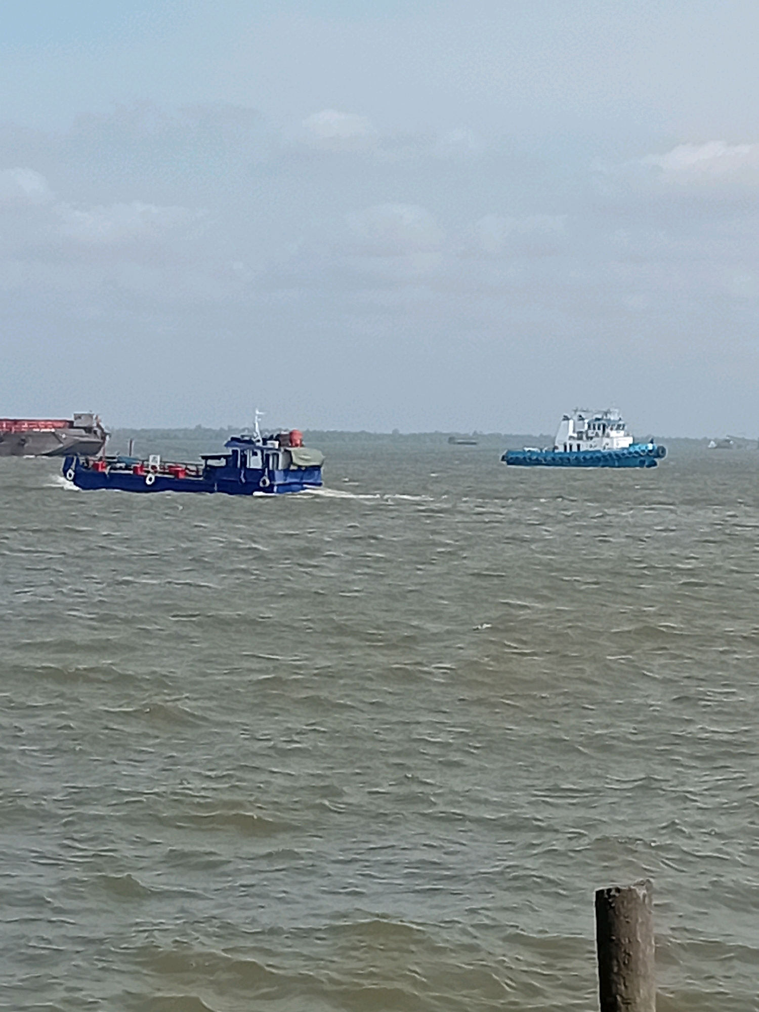 Two boats are sailing on a body of water. One boat is blue and has a white cabin. It is being towed by another larger boat that is dark gray with red accents. The second boat is a tugboat and is white with a blue stripe along its hull. The sky is cloudy and the water is choppy. The scene is located in Sungai Meriam, Indonesia. A wooden post is visible in the foreground.