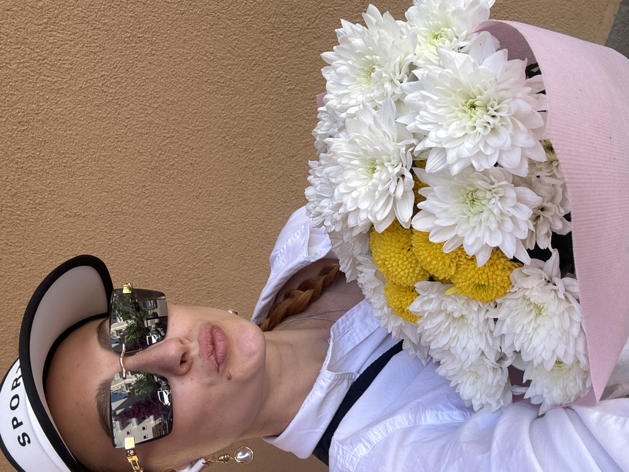 A light-skinned woman wearing a white sports visor, sunglasses, and a white button-down shirt holds a bouquet of white and yellow flowers wrapped in pink paper. A brown wall is visible in the background. The sunglasses reflect a building and trees, possibly located in an urban setting. A gold-colored earring hangs from her ear. She is positioned indoors or outdoors against the wall.