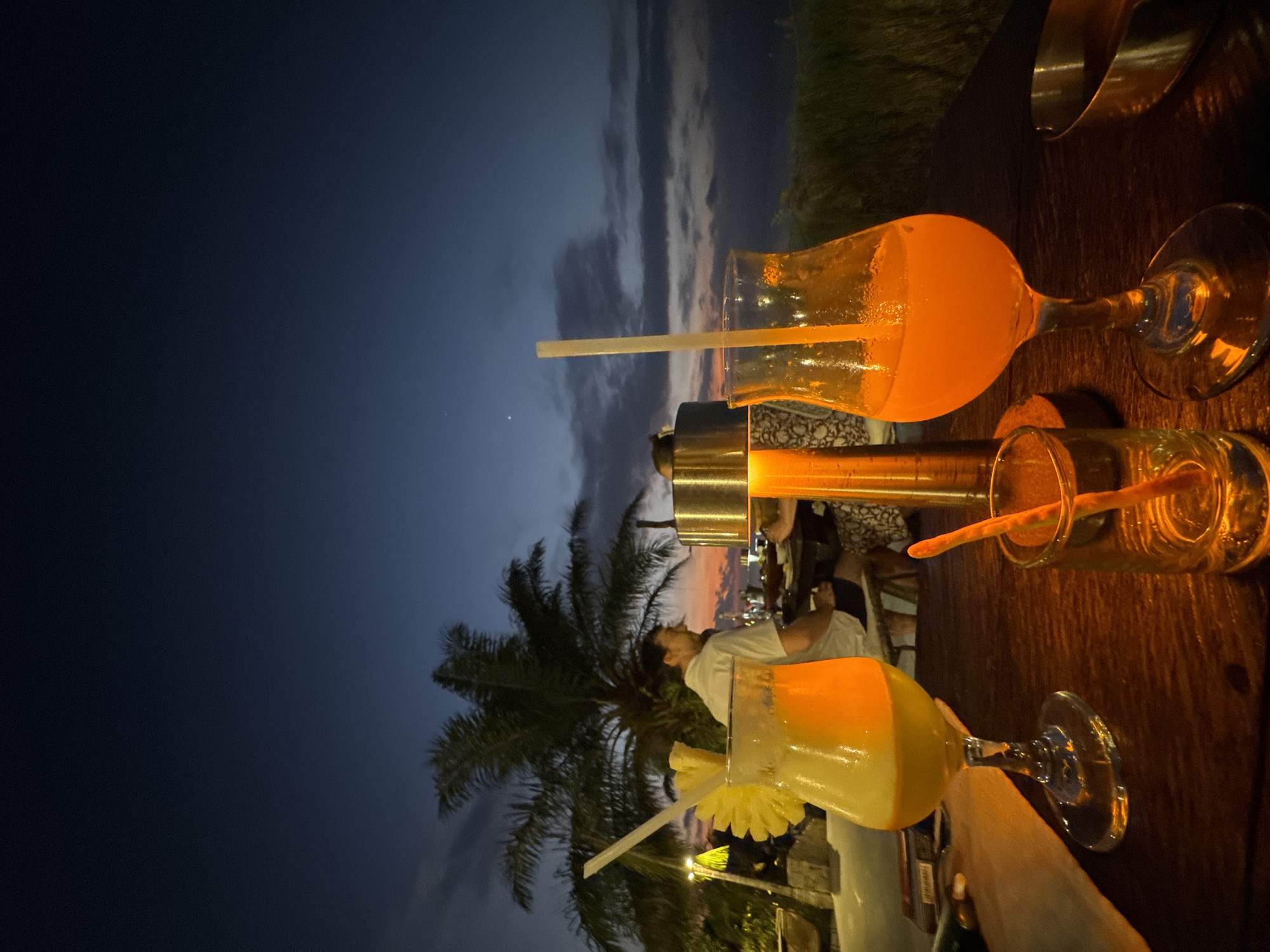 This image depicts an outdoor evening scene, likely at a bar or restaurant in Kuta Selatan, Indonesia. The setting appears to be a tropical location, characterized by visible palm trees.
In the foreground, a dark wooden table holds multiple drinks. Three main beverages are visible: two in stemmed glasses and one in a shot-glass-like container, all containing vibrant orange and yellow liquid that glows brightly from artificial light. One tall glass holds an orange drink with a white straw, while a wider glass contains a layered yellow and orange drink, garnished with a pineapple slice and another straw. A smaller glass also holds an orange drink with a short straw. A tall, metallic cylindrical object, possibly a table lamp or a heating/cooling unit, is positioned between the drinks.
Several people are present in the midground. A man wearing a white t-shirt is visible, looking upwards towards the sky and palm trees. Another individual is partially seen seated further back, behind the table.
The time of day is late dusk or early night, indicated by the sky transitioning from deep blue to lighter grey and orange hues at the horizon, with scattered clouds. The palm trees are silhouetted against this twilight sky, enhancing the tropical ambiance. The overall atmosphere is casual and relaxed, suggesting people are enjoying drinks in a pleasant outdoor environment. There is no visible text in the image.