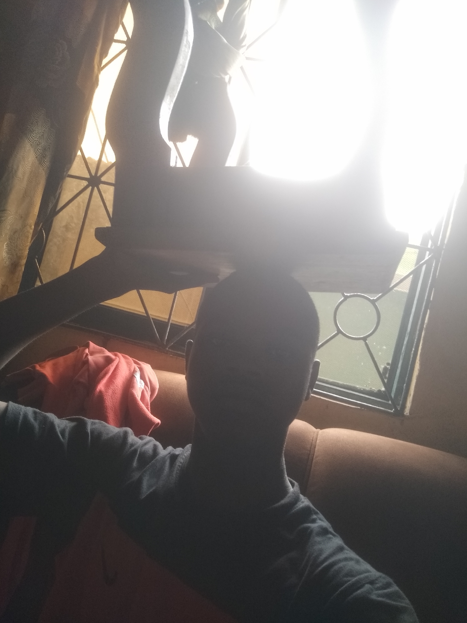 A person is depicted indoors, primarily in silhouette, holding a large, intensely bright, rectangular light source directly above their head. The light source is overexposed to pure white, obscuring its details. The person, seen from behind and slightly to the side, is wearing a dark, long-sleeved shirt and appears to be sitting on a tan or brown upholstered sofa.
Behind the person, a window is visible with a decorative metal security grille featuring geometric patterns including triangles and circles. Warm, natural light enters through this window. To the left of the window, a patterned curtain is partially drawn. On the sofa, near the window, a crumpled piece of red fabric, possibly a towel or blanket, rests.
The image is characterized by strong backlighting, causing the person and most interior elements to be heavily underexposed or silhouetted against the bright light source and the window. No text is visible in the image.