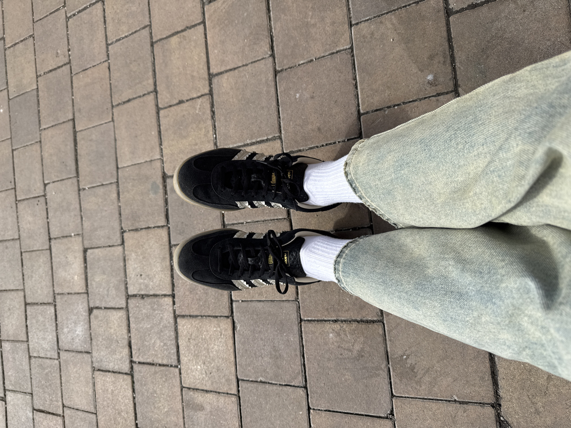 The image shows a view from above of a person's feet on a paved surface. The person is wearing black Adidas Samba sneakers with white stripes and white socks. The person is also wearing light blue denim jeans. The paved surface appears to be made of rectangular bricks. The setting might be somewhere in a city with such brick paving. The image is not sufficient to determine any further details about the location or the activities taking place. There is no evidence from the image to support the claim that the location is in the city of Moscow, Russia.