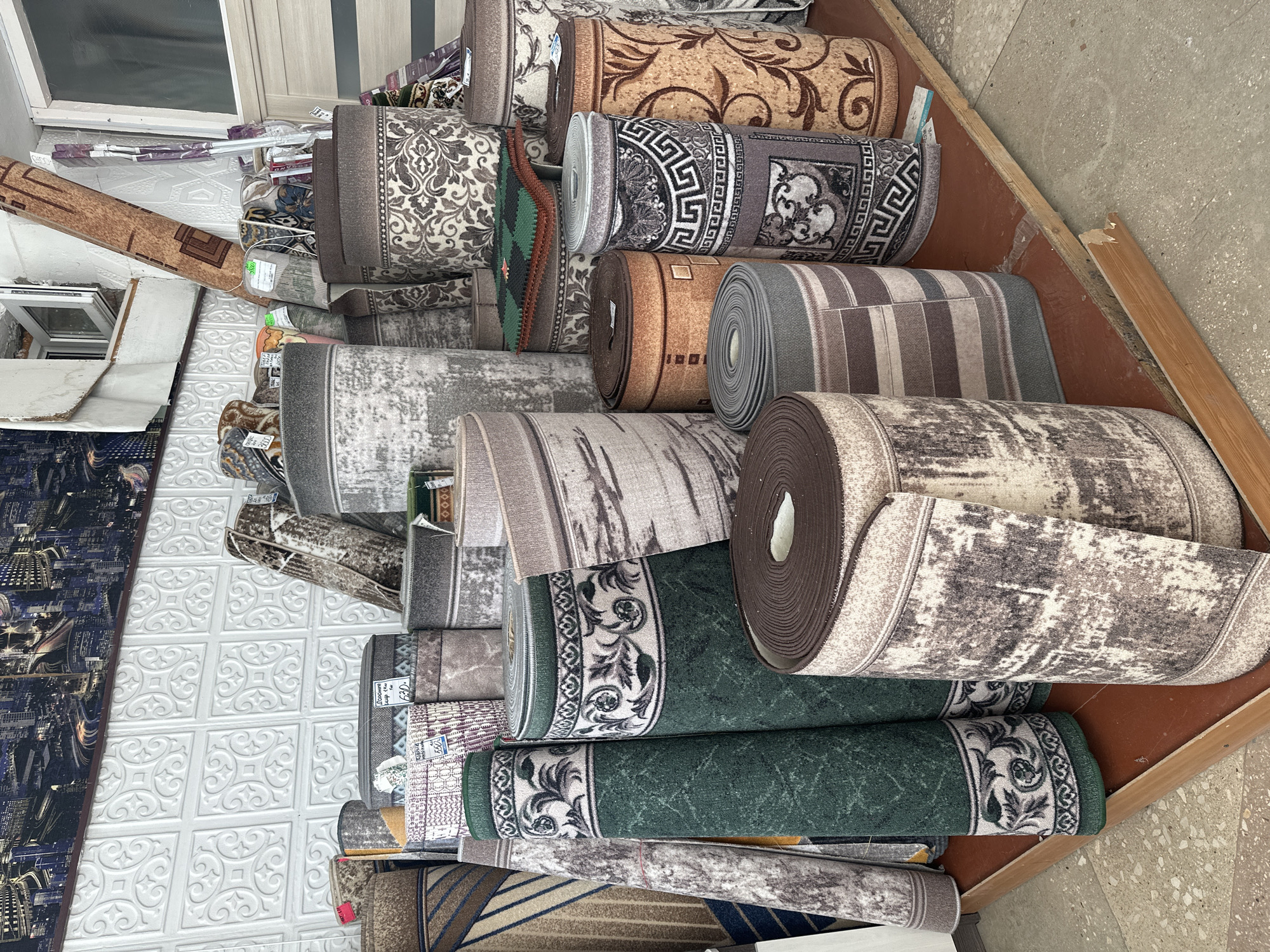 The image shows a close-up view of several rolled-up carpets displayed in a retail setting. The carpets are stacked on top of each other, with some leaning against a wall. The patterns on the carpets vary, including floral designs, geometric shapes, and abstract textures in shades of brown, gray, beige, and green. Some carpets have visible labels or price tags. The background includes part of a white tiled wall and a wooden floor. The lighting appears to be artificial, suggesting an indoor environment. The scene is focused on the merchandise, with no people visible. The overall impression is that of a carpet store or warehouse. There is no indication of the time of day or weather conditions.