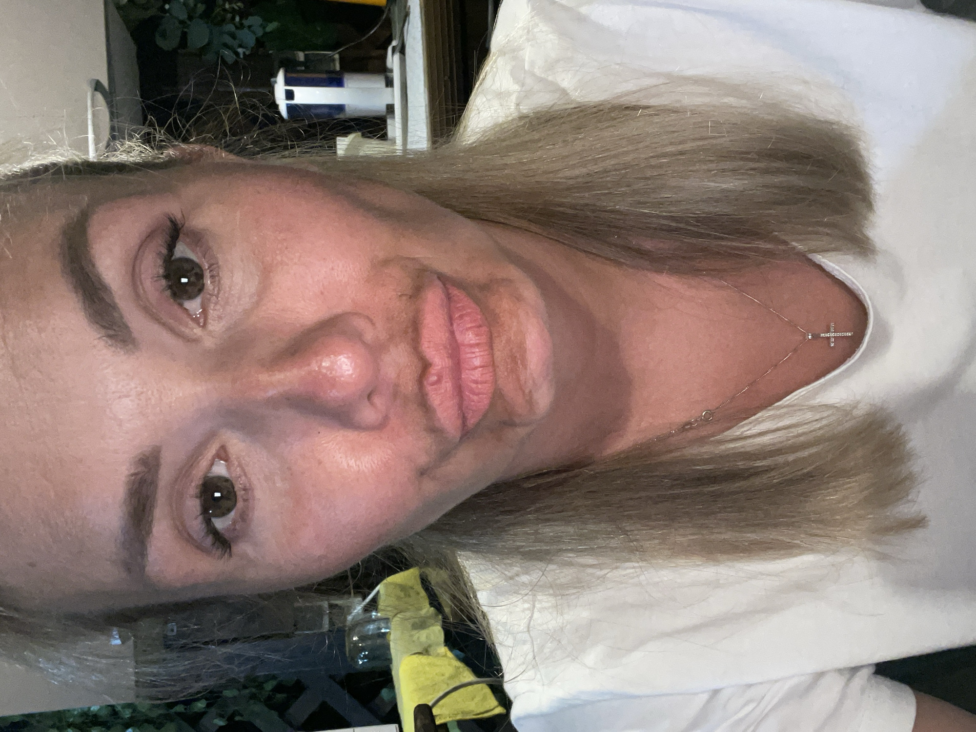 An indoor selfie shows a blonde woman with fair skin, wearing a white t-shirt and a silver cross necklace. She has brown eyes, well-defined eyebrows, and her lips are pursed. There is a smudge of brown makeup on her upper lip and chin. Her hair is blonde and shoulder-length. The background is blurred and appears to be an indoor setting with some objects and greenery visible. The lighting is somewhat dim. The location is unknown.
