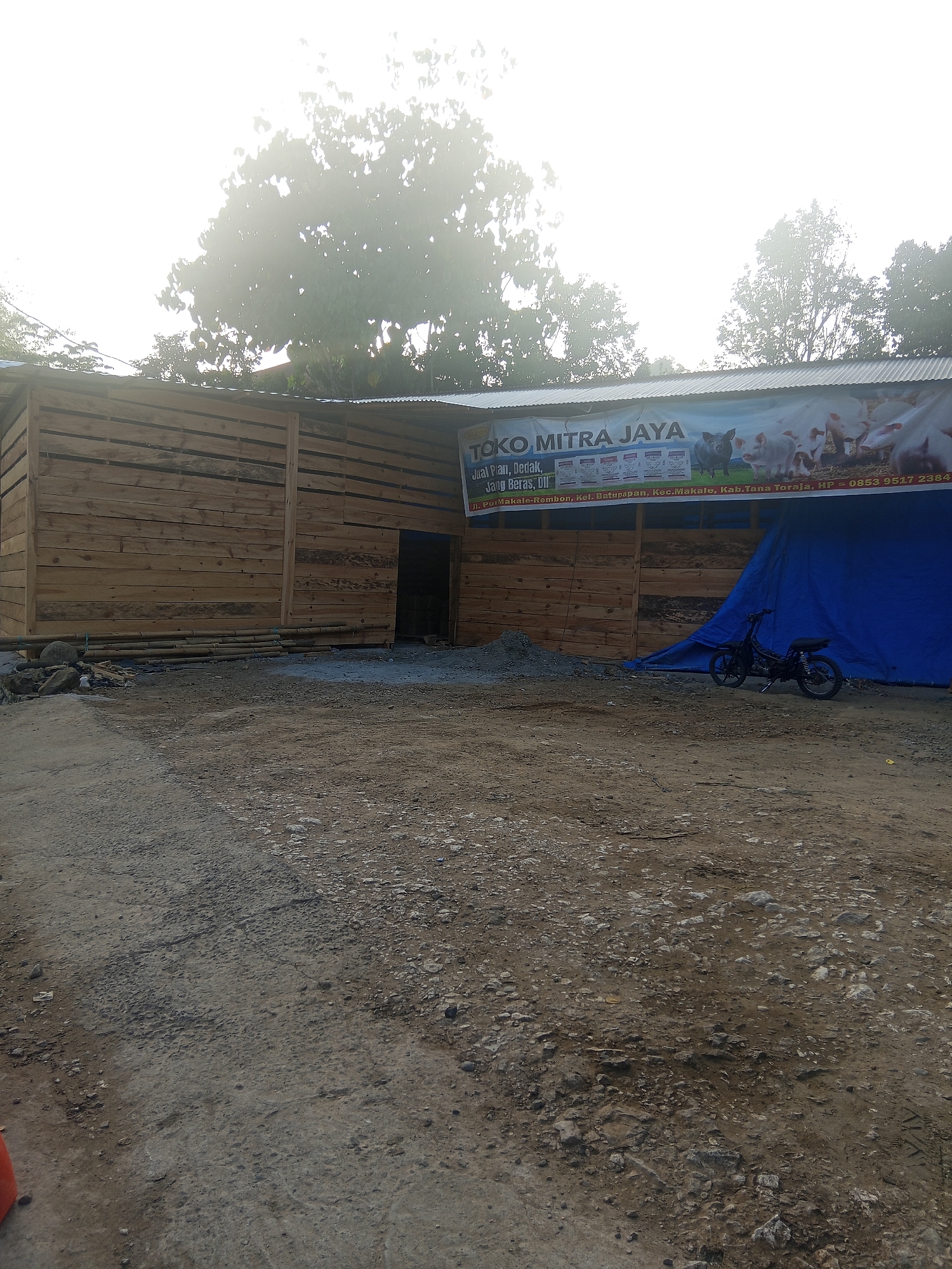 The image shows a store named "TOKO MITRA JAYA" located in Batu Papan, Indonesia. The store appears to be under construction or recently built, with a wooden exterior and a corrugated metal roof. A large blue tarp is spread out in front of the store, covering some of the area. A black motorcycle is parked near the tarp. The store's banner advertises "Jual Pakan, Dedak, Jagung Beras, dll" which translates to "Selling Feed, Bran, Corn Rice, etc.". The banner also features images of pigs. The surrounding environment is dusty and appears to be a rural or semi-urban area with trees in the background. The sky is hazy, suggesting it might be early morning or a misty day. There are no people visible in the image.