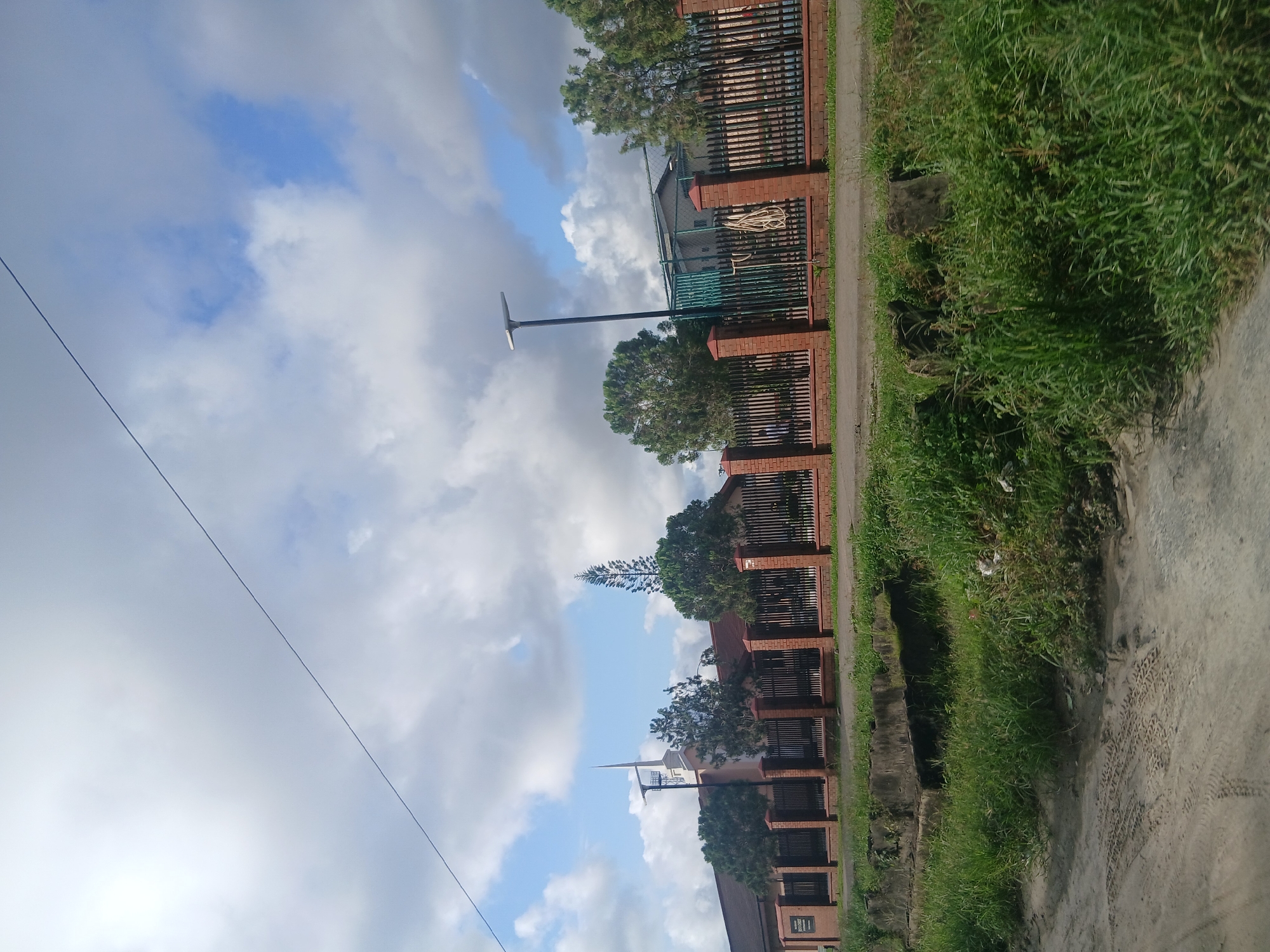 The image shows a street scene in Satellite Town, Nigeria, featuring a building that appears to be a place of worship, possibly a church, with a distinct spire. The building is partially visible behind a brick and metal fence. Trees are scattered along the property line. A street light is visible in the foreground. The sky is partly cloudy with a thin power line stretching across it. In front of the fence, there is a drainage ditch with overgrown vegetation. The foreground shows a dirt road with tire tracks.