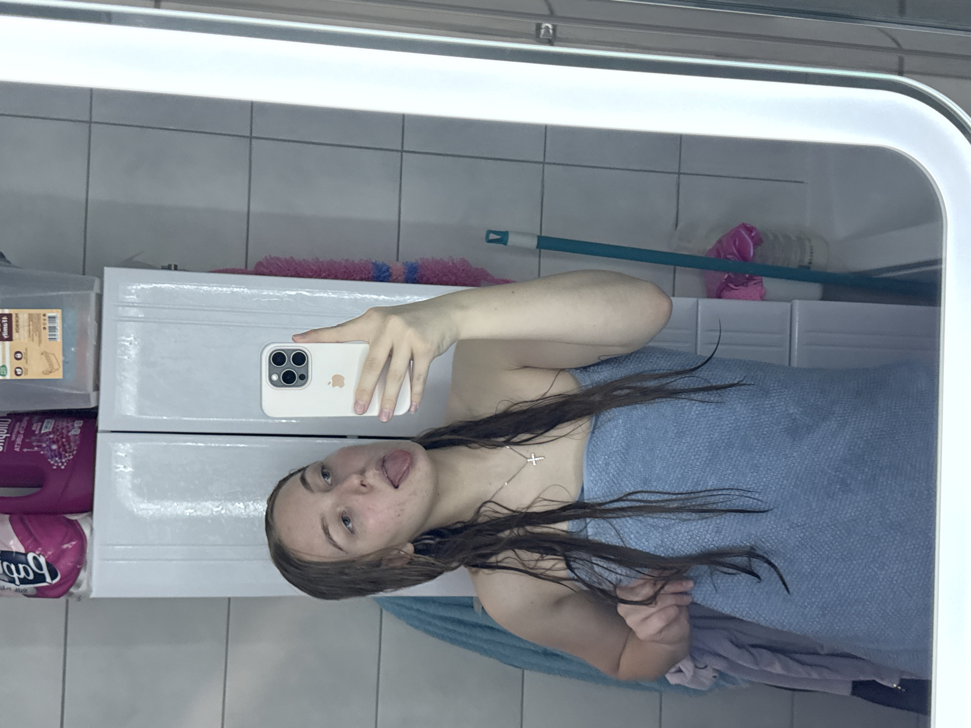 A young female, appearing to be in her late teens or early twenties, is depicted in a mirror selfie taken within a bathroom in Perm, Russia. Her long, wet, light brown hair hangs loosely around her shoulders. She is wrapped in a light blue textured towel, exposing her bare shoulders. Visible acne or redness is present on her cheeks. She wears a silver-colored chain necklace with a small cross pendant. Her expression is playful, with her tongue extended, and her gaze directed towards her left. She holds a light-colored smartphone with a triple-lens camera in her right hand to capture the image. Her left hand is partially visible, holding the towel.
The background, a reflection in the mirror, reveals a tiled bathroom environment. The walls are covered in light grey/white rectangular tiles with visible grout lines. Above a counter or sink area, two white, paneled cabinets are mounted. Various items are stored atop these cabinets, including a pink plastic bottle, a purple plastic bottle, and a clear plastic container with a brown label. A blue mop with a light-colored tip leans against the wall on the right side of the frame. A pink scrubbing brush or sponge is suspended on the wall between the cabinets. To the left of the female, a blue towel hangs, and a pile of light-colored textiles (possibly clothing or towels) rests on a surface. The mirror itself has a wide, illuminated frame on its right edge, suggesting integrated lighting. The bathroom appears orderly.