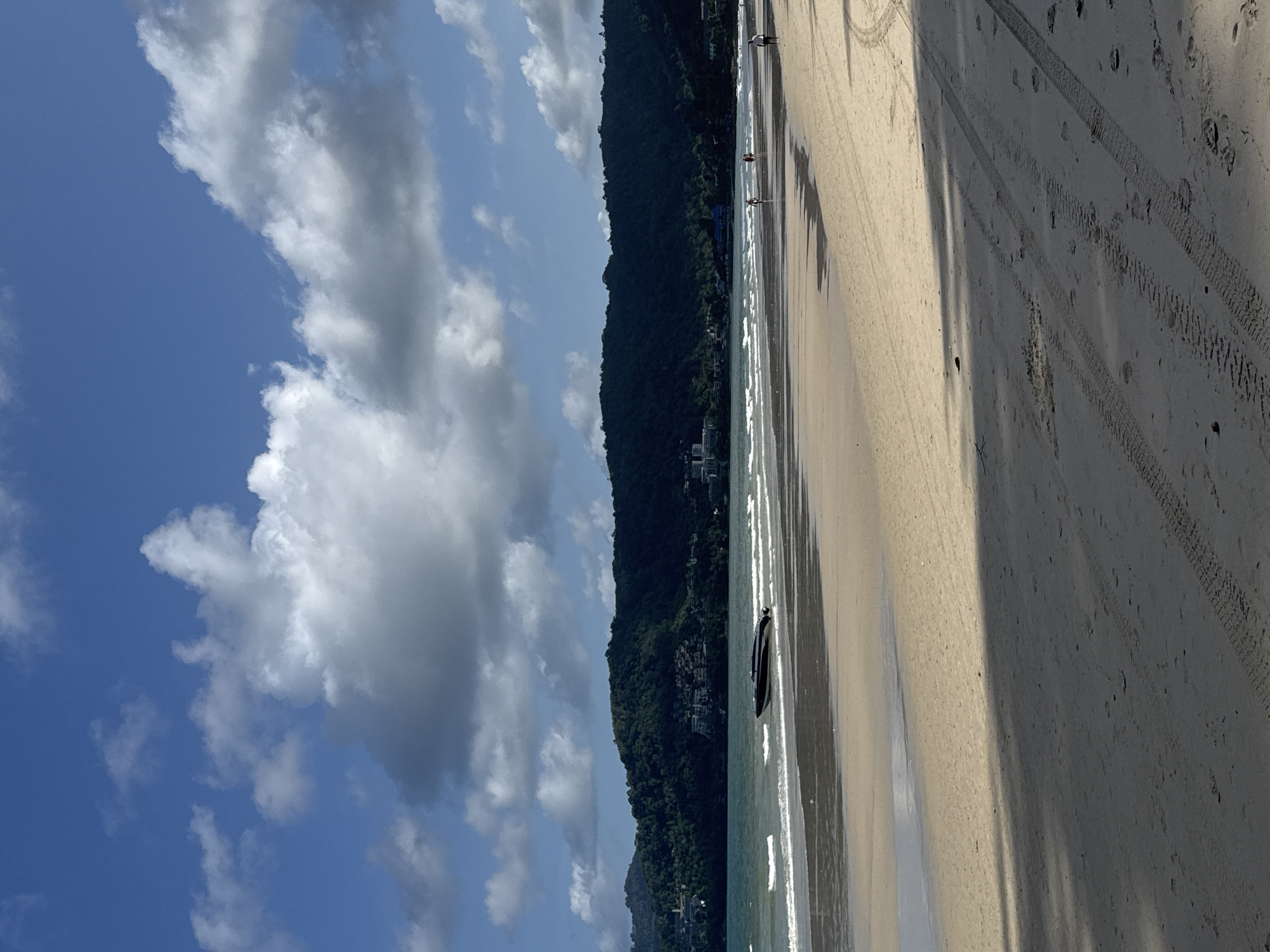 The image depicts a beach scene in Patong, Thailand, under a partially cloudy blue sky. In the foreground, a sandy beach shows tire tracks and scattered debris, with the dark shadow of a palm tree extending across it. The shallow water near the shore has gentle waves breaking on the sand. Further out, a black speed boat is partially beached in the shallow water. In the background, a lush green, tree-covered hill with scattered buildings rises above the water, forming the coastline. Several people are visible walking on the beach in the middle ground, some near the water's edge.