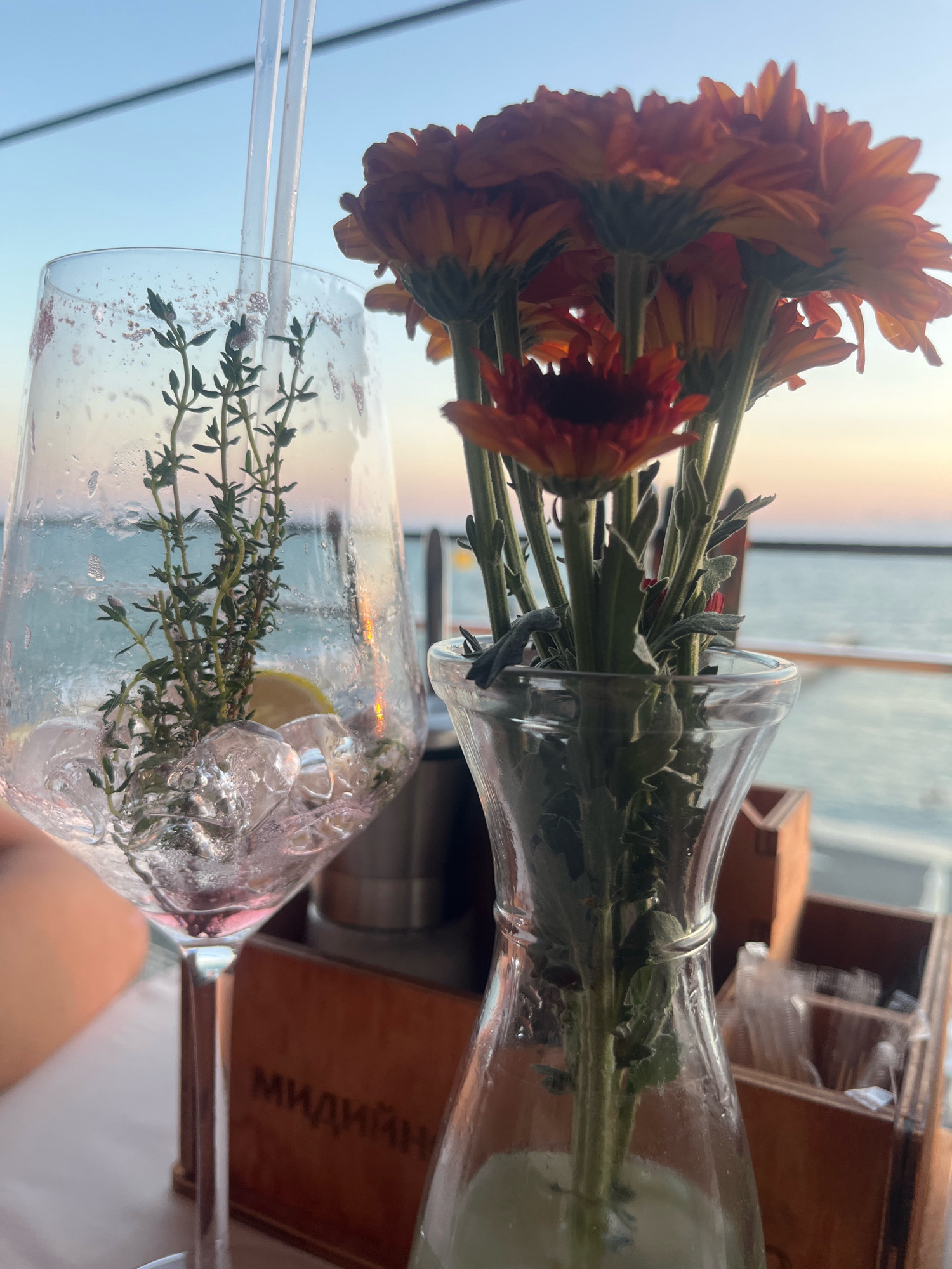 The image shows a close-up of a table setting with a drink and flowers, likely at an outdoor cafe or restaurant in Sochi, Russia, given the style of the drink and the presence of a wooden crate with Cyrillic lettering. In the foreground, a tall glass with a pink hue, filled with ice, water, a sprig of thyme, and a slice of lemon, has two clear straws inserted. Beside the glass, a small glass vase contains a bouquet of orange chrysanthemums. Behind these items, a wooden crate with the partially visible Cyrillic word "МИД" and other letters is situated. The background reveals a blurred body of water, likely the Black Sea, with a hazy sky suggesting either sunrise or sunset. The overall scene evokes a relaxed, possibly vacation setting.