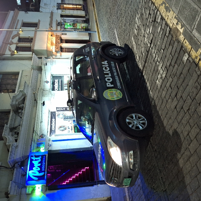 A Peruvian police car is parked on a cobblestone street next to a building with a nightclub called "Port Discoteca." The club's sign glows with blue light, and the entrance features pink neon. Banners with "Grékos" are visible above. The police car has "POLICIA" and "COMISARIA PALACIO VIEJO" printed on it. The street is lit by streetlights and building lights.