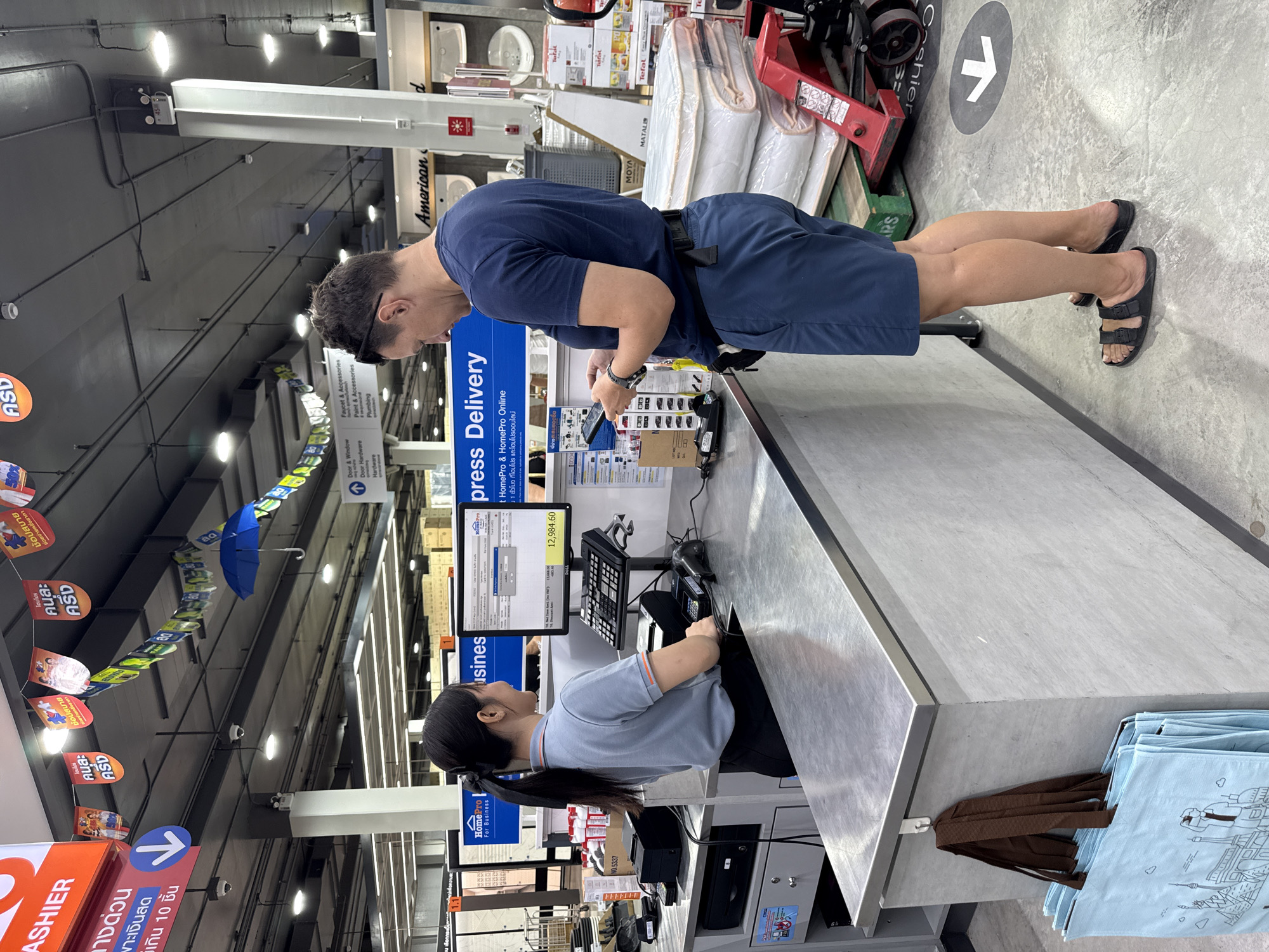In a HomePro store in Baan Chaweng Noi, Thailand, a male customer wearing a navy blue t-shirt and navy blue shorts stands at a checkout counter. He is holding a smartphone in his left hand and appears to be looking at it. A female cashier, wearing a light blue polo shirt with orange trim and black pants, stands behind the counter and is operating the point-of-sale system. The counter is made of brushed metal and has a computer monitor displaying a transaction summary, a cash register, a credit card terminal, and a barcode scanner. Several shopping bags are hanging from a hook on the side of the counter. In the background, shelves are stocked with various merchandise, including mattresses and building materials. Overhead, decorative banners and umbrellas hang from the ceiling. The floor is concrete, with directional arrows indicating pathways.