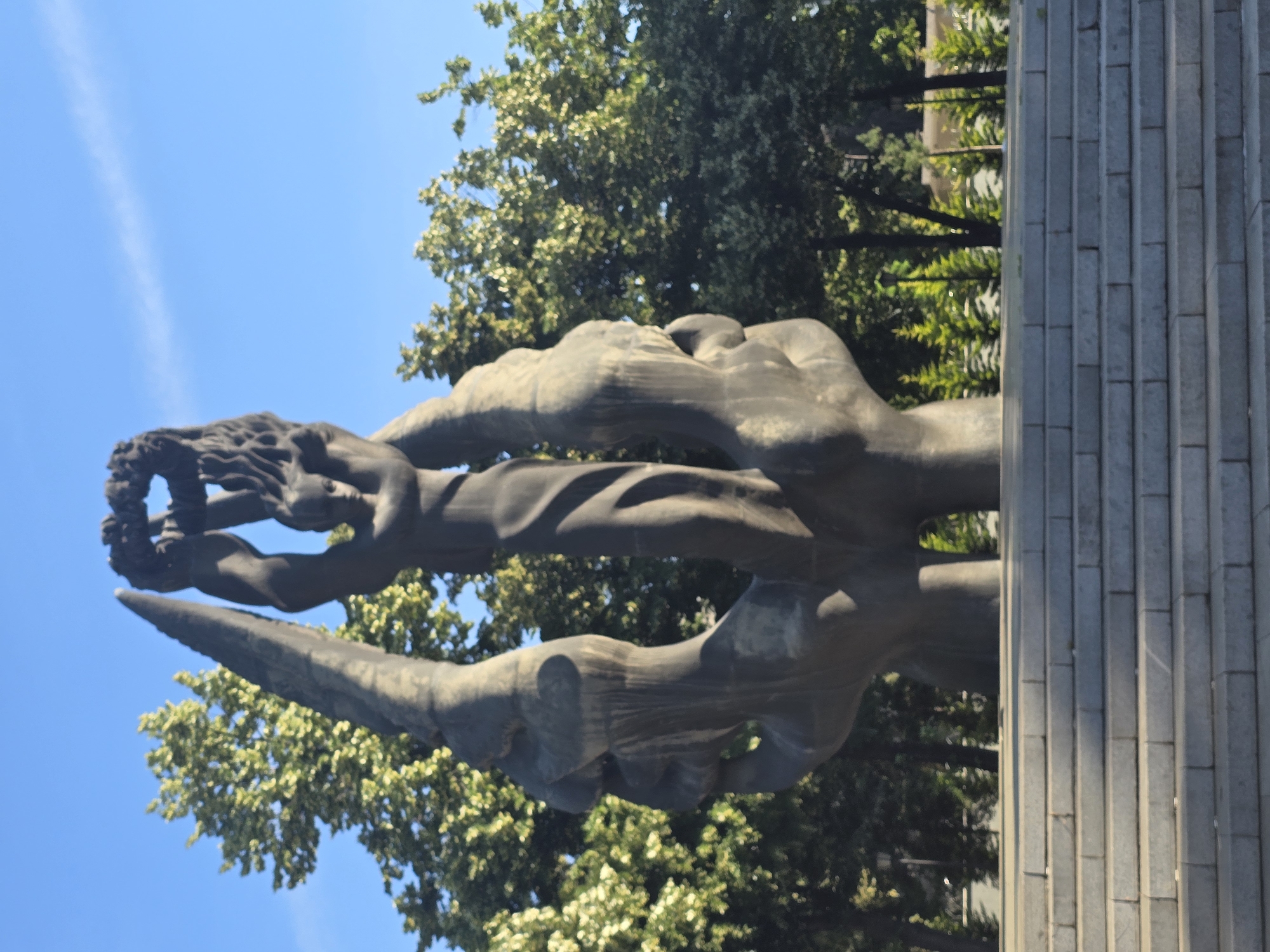 The image shows a large, dark-colored sculpture depicting a human figure holding a wreath above its head, supported by two large, abstract hands or wing-like structures. The sculpture is made of stone or concrete and stands on a stone base, with a wide set of stone stairs leading up to it. The background consists of lush green trees and a clear blue sky with a few wispy clouds. The scene appears to be outdoors, possibly in a park or public square, and is bathed in bright sunlight, casting shadows on the stairs. There are no people visible in the image.