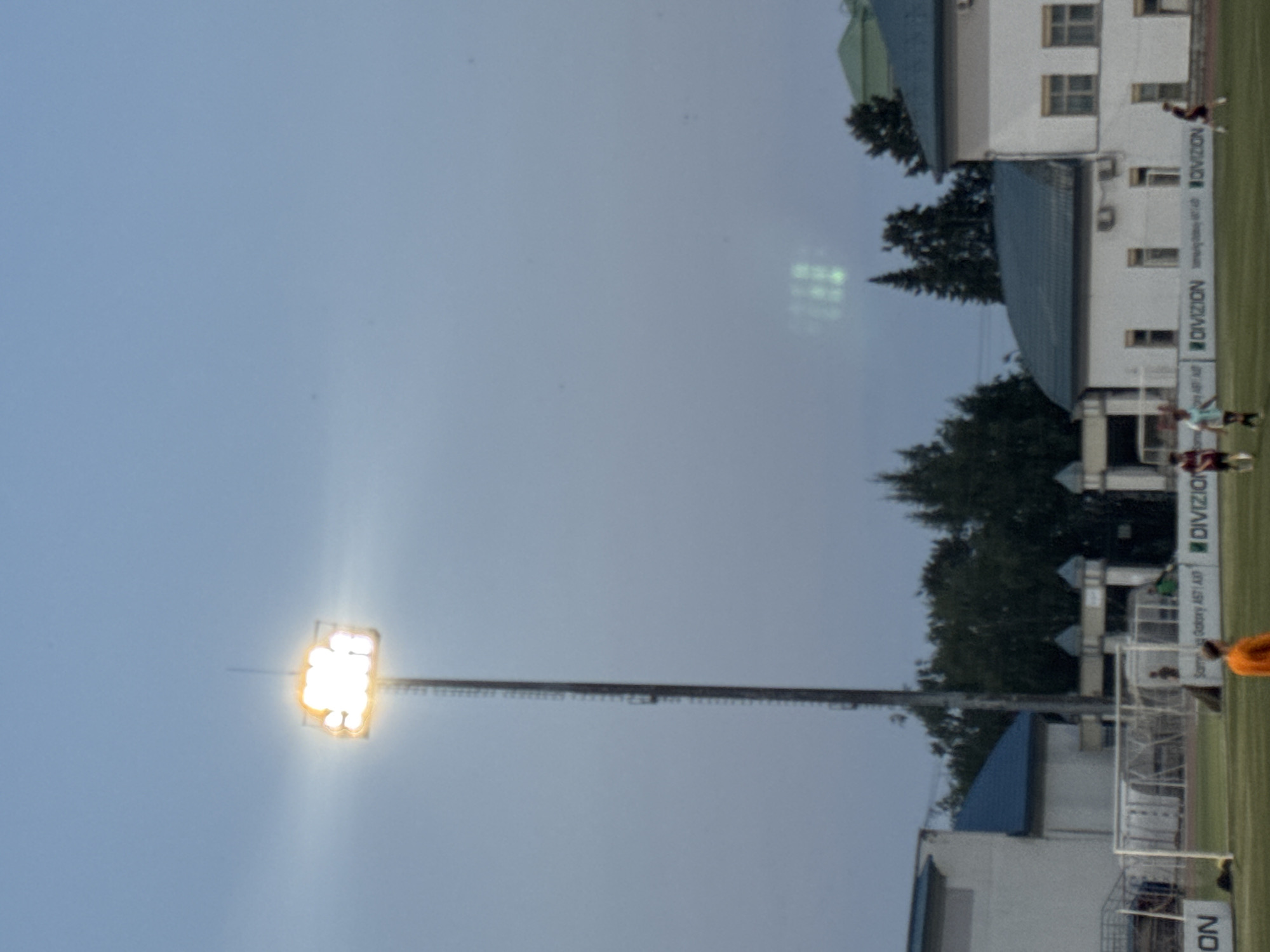 The image depicts an outdoor sports field, likely a soccer stadium, at dusk or during twilight. A tall, industrial-looking floodlight tower is prominent on the left side of the frame, its bright light illuminating a portion of the sky and the field. In the background, there are several buildings with dark blue, sloped roofs and white walls. Some of these buildings appear to be sports facilities or administrative buildings associated with the stadium.
On the field itself, there are a few individuals, some of whom appear to be players, engaged in activities. One person in an orange shirt is walking on the field. Another individual in a light blue and white jersey is running. A person in a dark red jersey is also visible running. There are soccer goals present on the field, suggesting a game or practice is underway or has recently concluded. Advertising banners with text such as "DIVISION" and "Samsung Galaxy A51 A5P" are affixed to the fence along the edge of the field.
The sky is a muted blue-grey, indicating the transition from day to night. There is a slight haziness or mist in the air, particularly noticeable in the upper right quadrant where some faint, greenish light patterns are visible, possibly a reflection or lens flare. Trees are visible in the background, contributing to the outdoor setting.
While the image provides a visual depiction of a sports venue and activities, it does not contain sufficient information to definitively identify the specific location within the city of Ufa, Russia. However, the presence of a soccer stadium, floodlights, and associated buildings are consistent with sports facilities found in many cities.