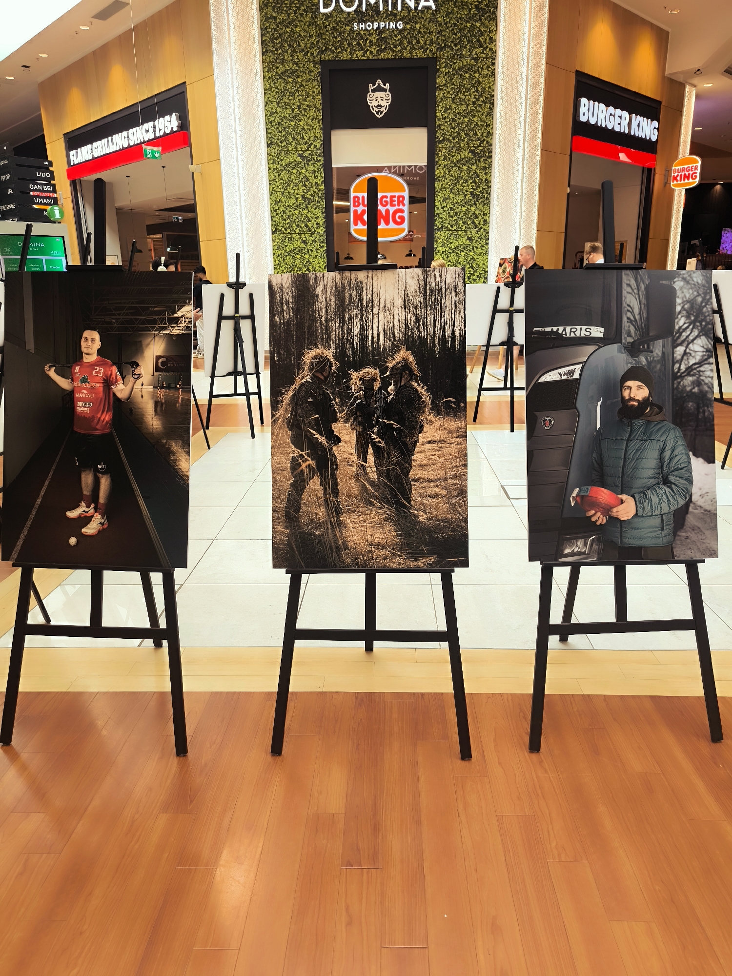 In the Domina Shopping mall in Riga, Latvia, three large photographs are displayed on black easels on a polished wooden floor. The leftmost photograph shows a man in a red t-shirt with the number 23 on it, wearing black shorts and athletic shoes, holding two controllers. He is in an indoor gymnasium. The central photograph depicts three people in camouflage gear, standing in tall, dry grass with trees in the background. The rightmost photograph features a man with a beard, wearing a beanie and a green jacket, holding a red strap in front of a truck. Behind the easels, store signs for "FLAME GRILLING SINCE 1954" and "BURGER KING" are visible, along with decorative green wall panels.