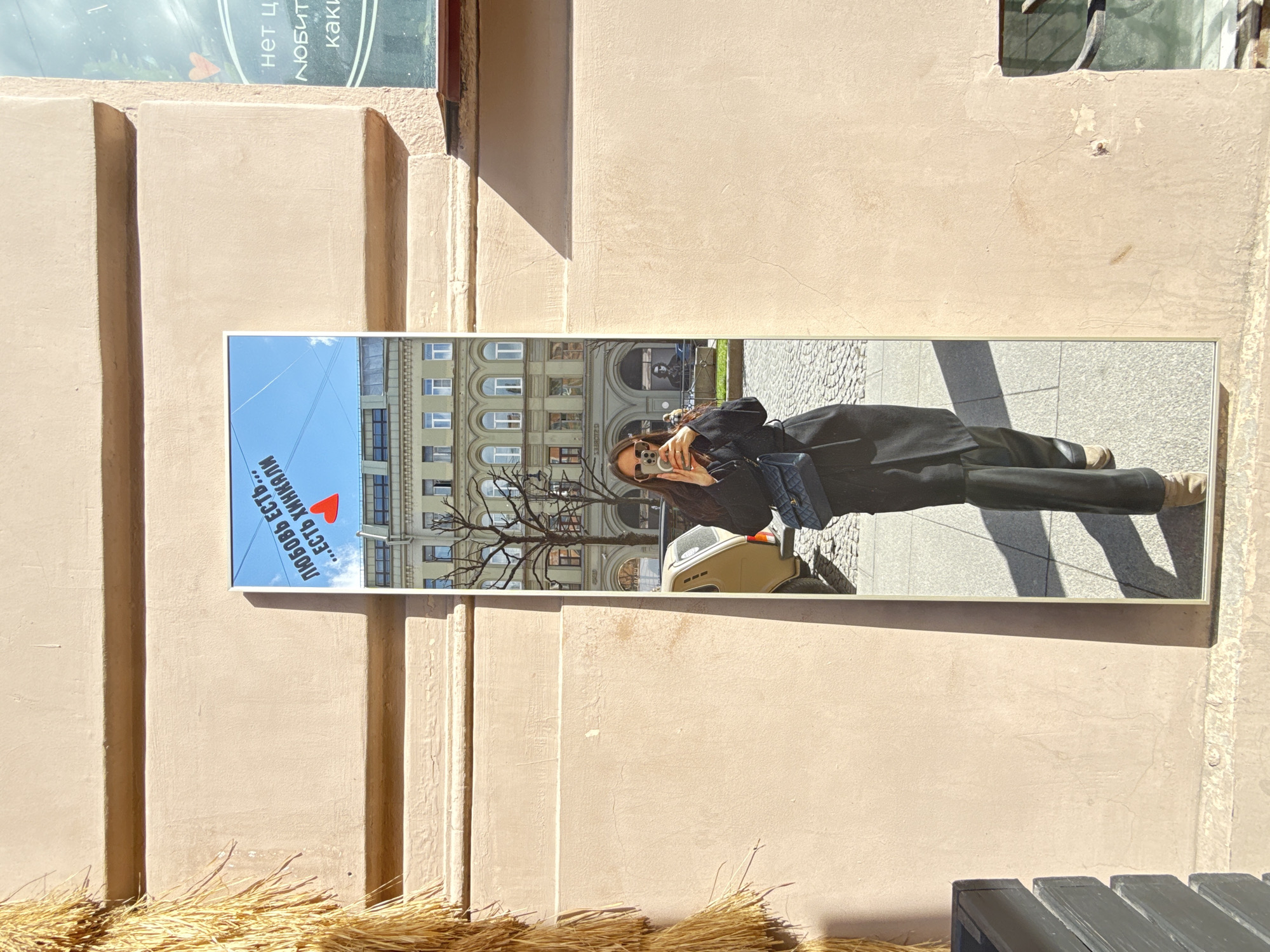 A mirror hangs on a light-colored wall with vertical indentations. The mirror reflects a woman taking a selfie with a building in the background. The reflected building's architecture indicates a location in Saint Petersburg, Russia. The woman is wearing a dark coat and sunglasses. A small vehicle is also visible in the reflection. The ground near the wall is covered with dry grass and a black bench. A sign is visible above the mirror, but only some words are readable, including "...есть хинкали...", which translates to "...there is khinkali..." and the word "любовь" which means "love".