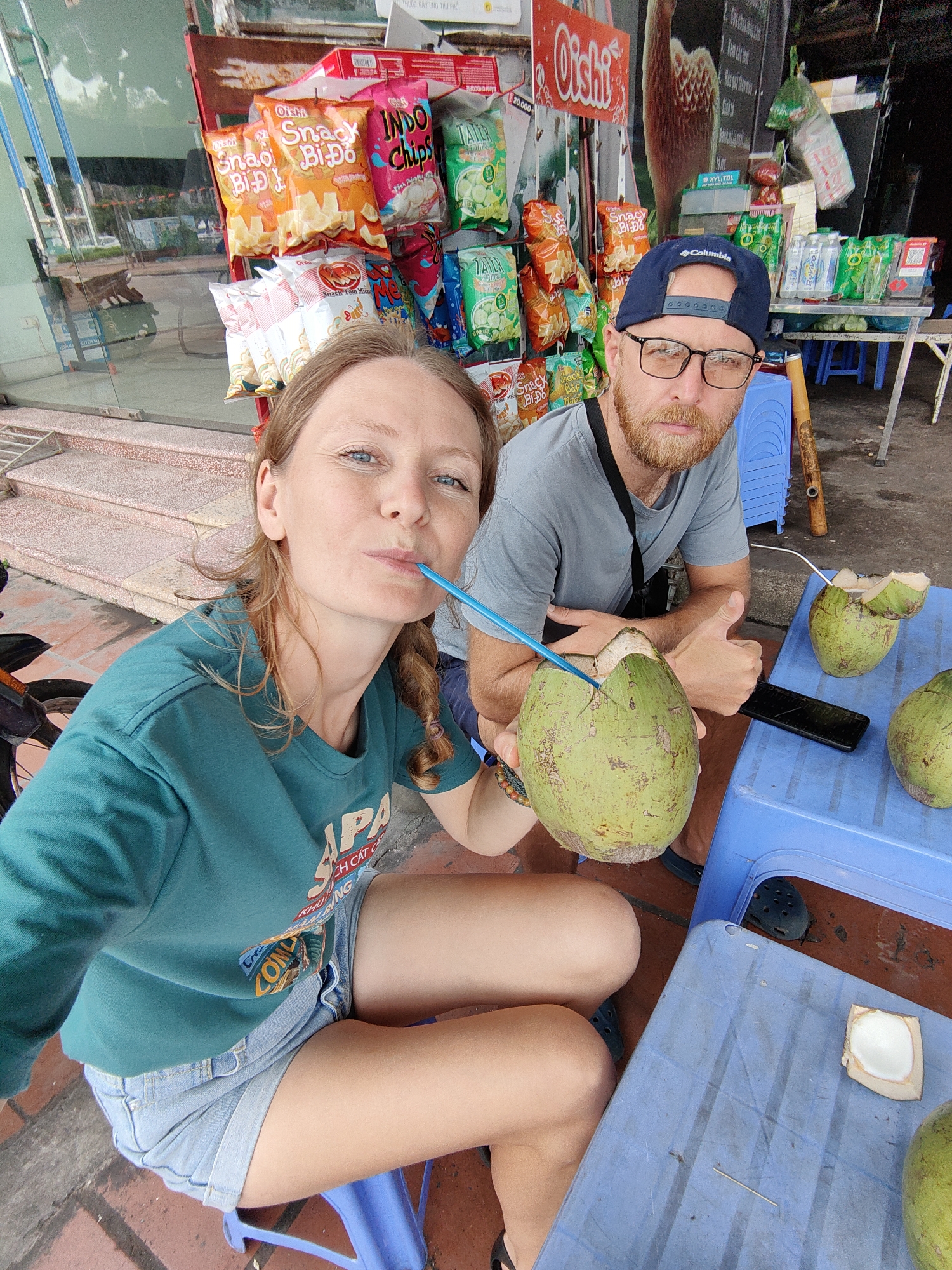 Two individuals are seated at a blue plastic table on a red-tiled surface, possibly a sidewalk, with a snack shop as a backdrop. A woman with braided hair and a green t-shirt is holding a green coconut with a blue straw inserted, and drinking from it. Beside her, a man with a beard, glasses, and a blue baseball cap gives a thumbs up.
On the table, there is a phone, two other coconuts, and a piece of cut coconut. The snack shop displays an array of packaged snacks, including chips with Vietnamese writing. Behind the man, there are stacks of blue plastic stools and green bottled beverages. The image captures an outdoor scene, suggesting a casual moment in a populated area. Based on the t-shirt "KHU NH CAT CA", this could be in Cao Xanh Ward, Vietnam.