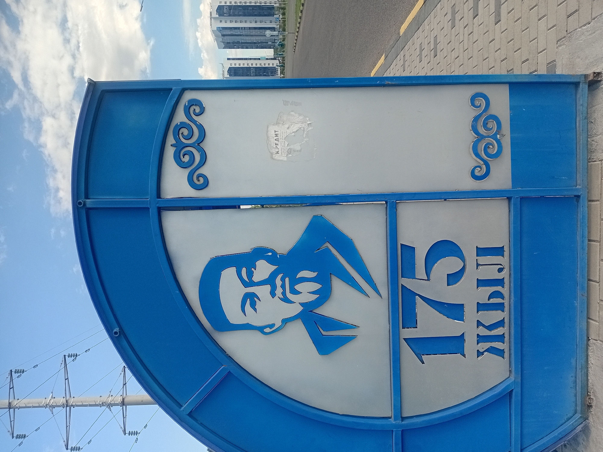 An outdoor medium shot captures a close-up of a blue metal gate with decorative elements and text. The gate features a stylized portrait of a man wearing a hat and a beard, rendered in blue against a silver background. Below the portrait, large blue numbers "175" are visible, followed by the Cyrillic text "ЖЫЛ", which translates to "years" in Kazakh. The gate has an arched top and is adorned with blue scrollwork on the right side. A torn white sticker with Cyrillic text, possibly an advertisement, is affixed to the silver panel on the right. In the background, a paved road runs alongside a sidewalk, with modern high-rise buildings in the distance under a partly cloudy blue sky. The scene appears to be located in the city of Semey, Kazakhstan, based on the text on the gate, which likely signifies an anniversary related to the city or a local institution.