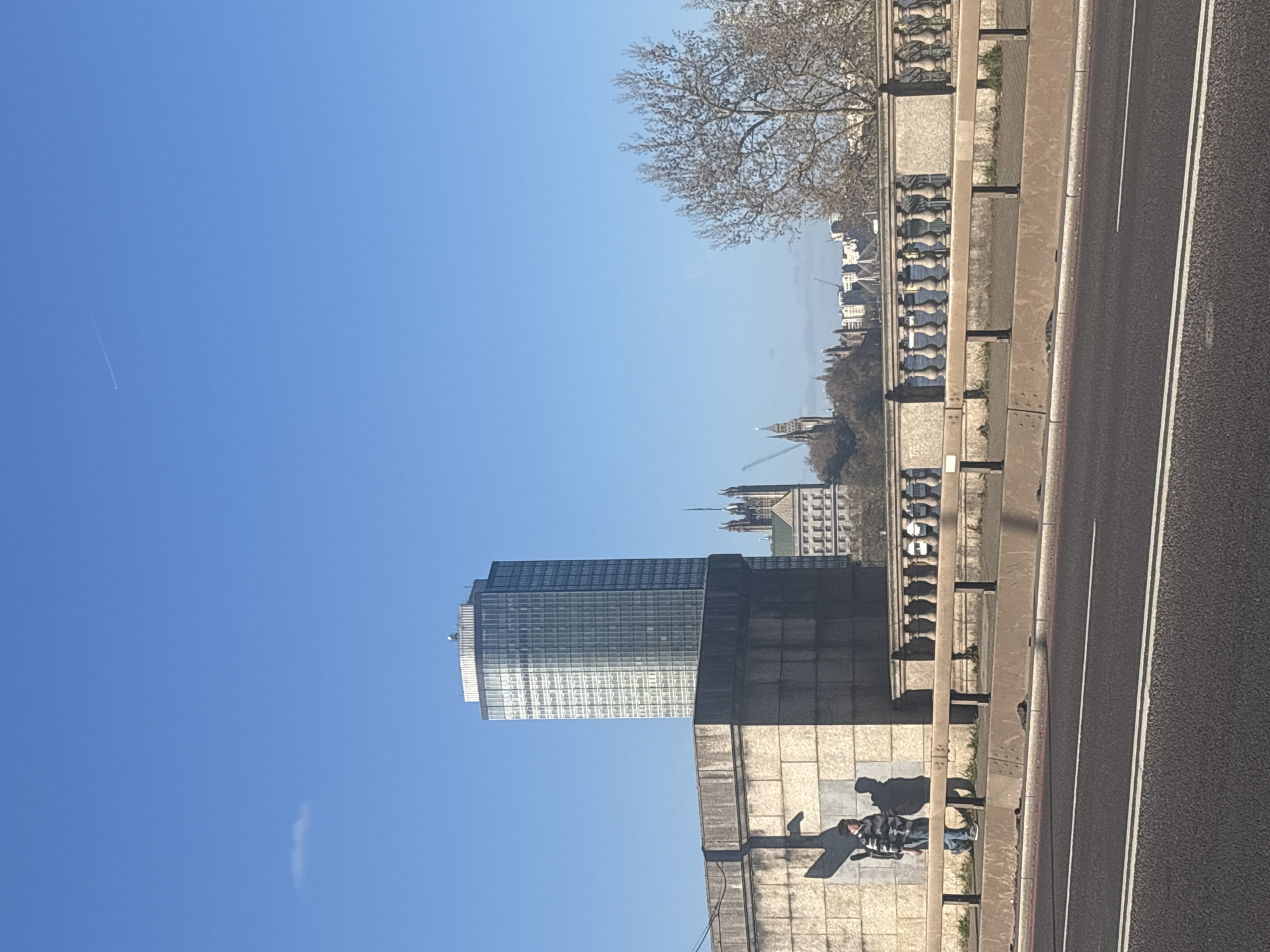 The image shows a view in London, United Kingdom. The Houses of Parliament and the Millbank Tower are visible. A person is lying on a wall next to a road. The sky is clear blue. A tree with bare branches is also in the frame.