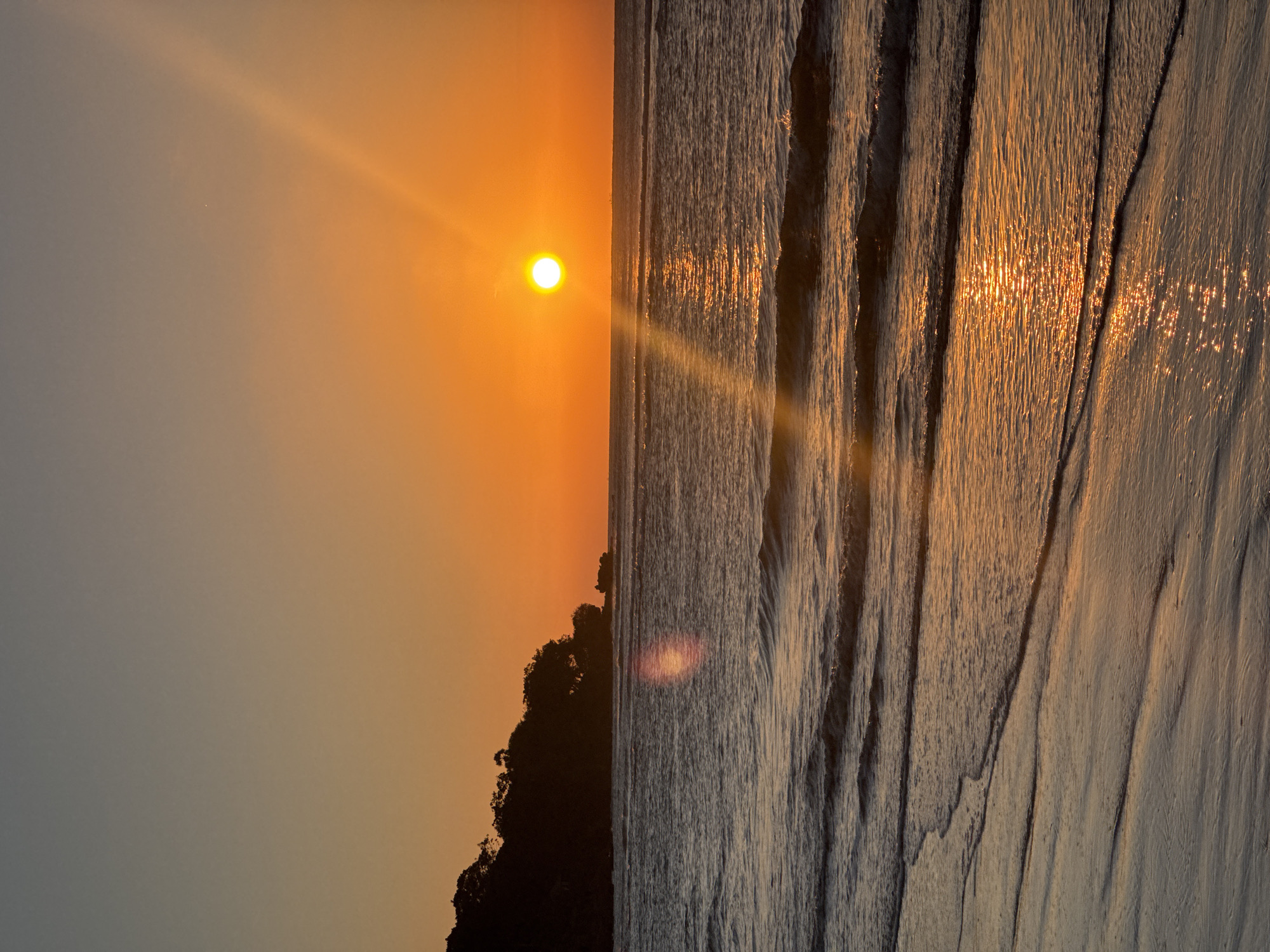 The image captures a serene coastal landscape during either sunrise or sunset in Kuta Selatan, Indonesia. The scene is dominated by warm, luminous colors.
In the upper-left portion, a brilliant, circular sun glows intensely with an orange-yellow light, emitting prominent sunbeams and a distinct lens flare that stretches across the sky. The sky itself displays a gradient, transitioning from vibrant orange and yellow immediately around the sun to a softer, greyish-blue hue towards the top left.
A dark, jagged silhouette of a landmass, likely a cliff or island with hints of vegetation at its crest, stretches across the middle of the frame, acting as a natural divider between the sky and the water below.
The lower half of the image features the ocean or a wet sandy beach. The water shows distinct parallel lines and textured patterns, indicative of receding waves or ripples on a shallow surface. The sun's golden light reflects brightly off the wet areas, creating shimmering streaks and highlights on the water's surface.
The overall atmosphere suggests a tranquil moment during the golden hour at a coastal location. There is no visible text in the image.