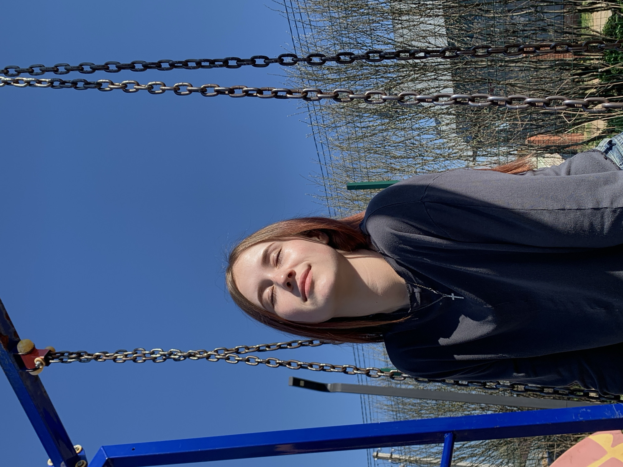 A young woman with brown hair is lying on a swing in a playground. She has her eyes closed and is smiling slightly. The swing is blue and has metal chains. The sky is blue and clear. There are trees in the background. The playground is in the city of Фоки, Russia.
