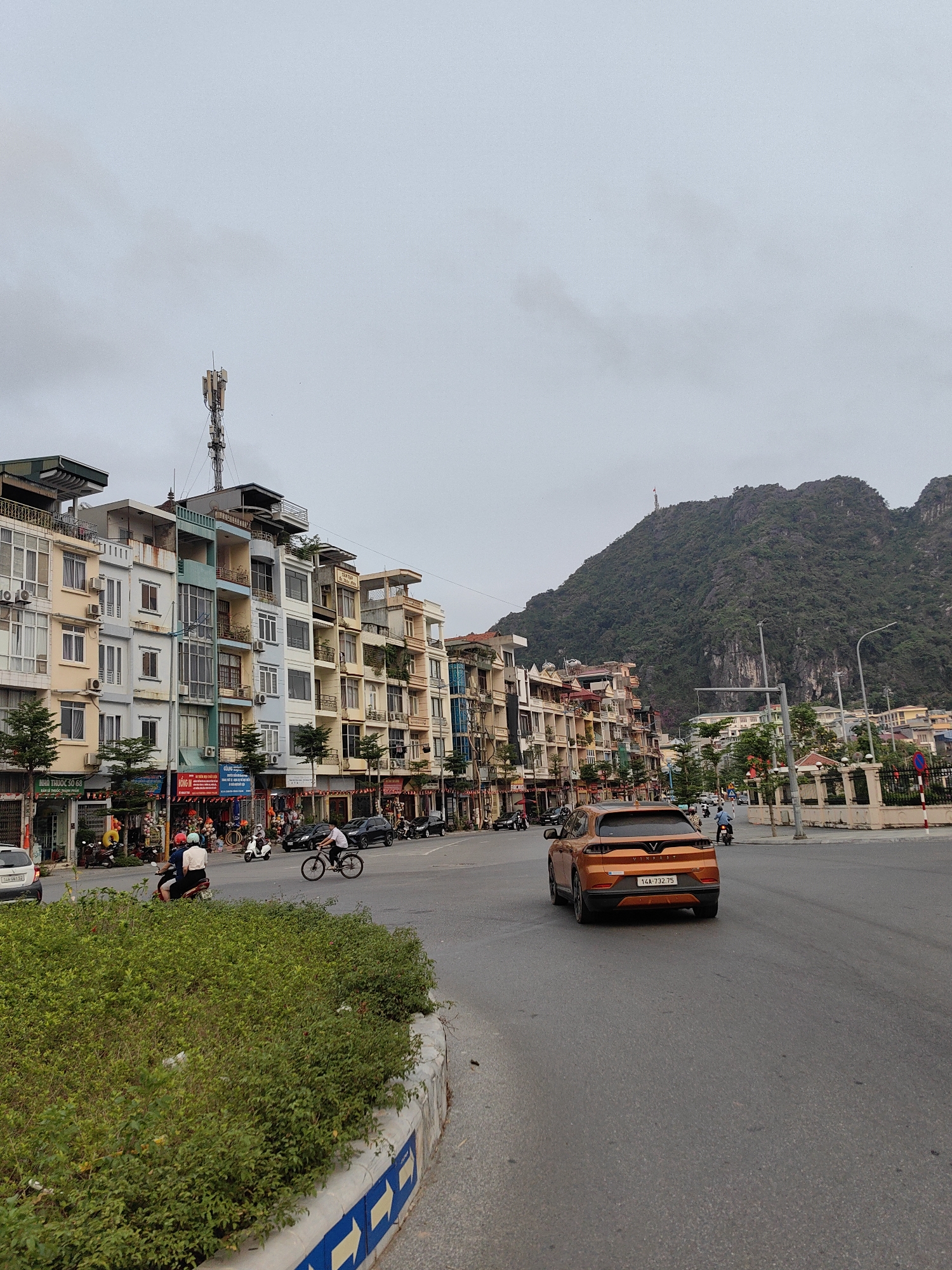 The image depicts a street in Hong Gai Ward, Vietnam. Along the left side of the street, there is a row of multi-story buildings with various architectural styles and colors. On the ground level, some of these buildings house businesses. The street scene includes pedestrians, a cyclist, motorbikes, and an orange car with the license plate 14A-732.75. In the background, a large, verdant mountain rises prominently. An antenna is visible atop one of the buildings. The sky is overcast. In the foreground, there is a landscaped area with greenery and a curb marked with directional arrows.